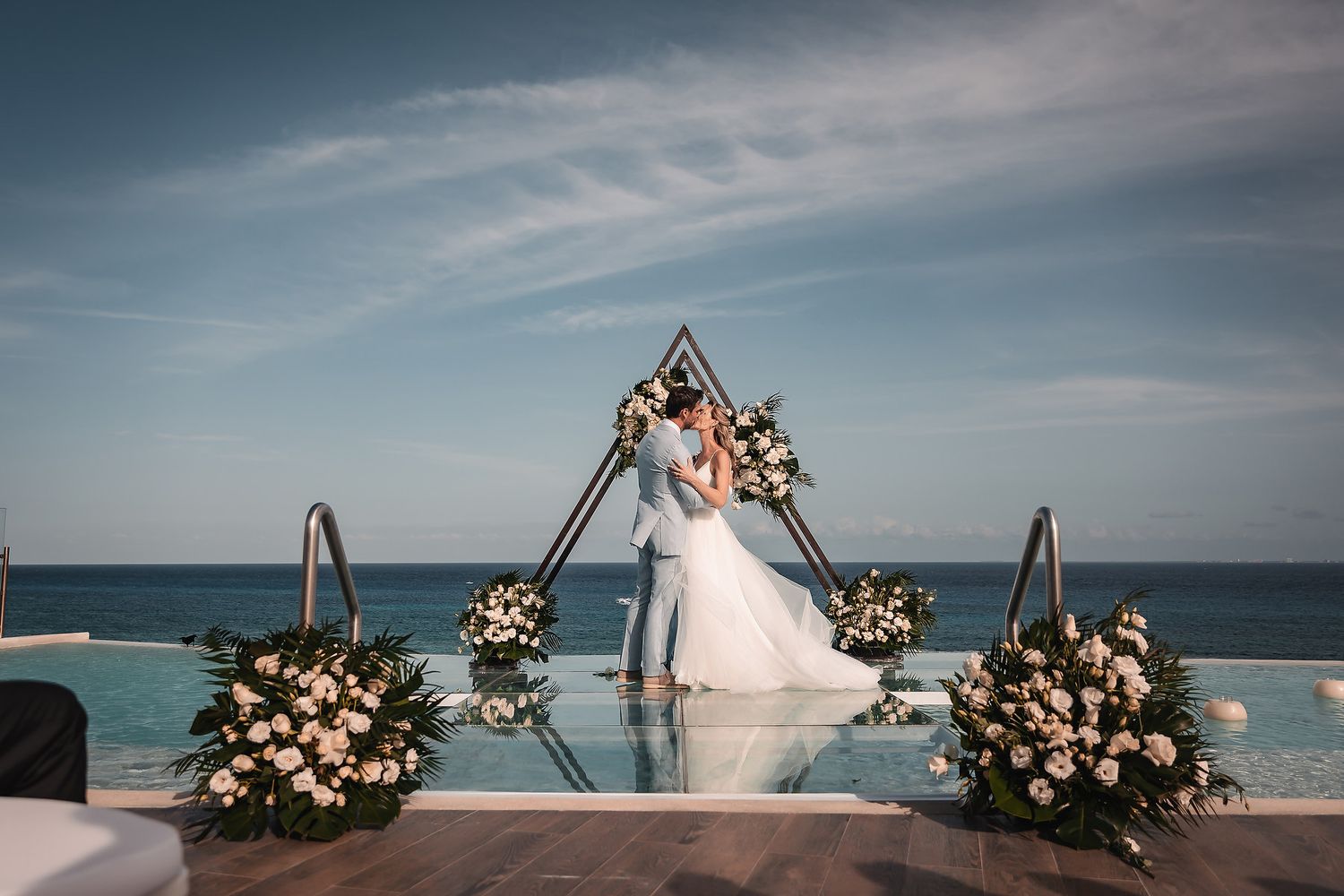 Wedding ceremony setup with triangular floral arch overlooking infinity pool and ocean.