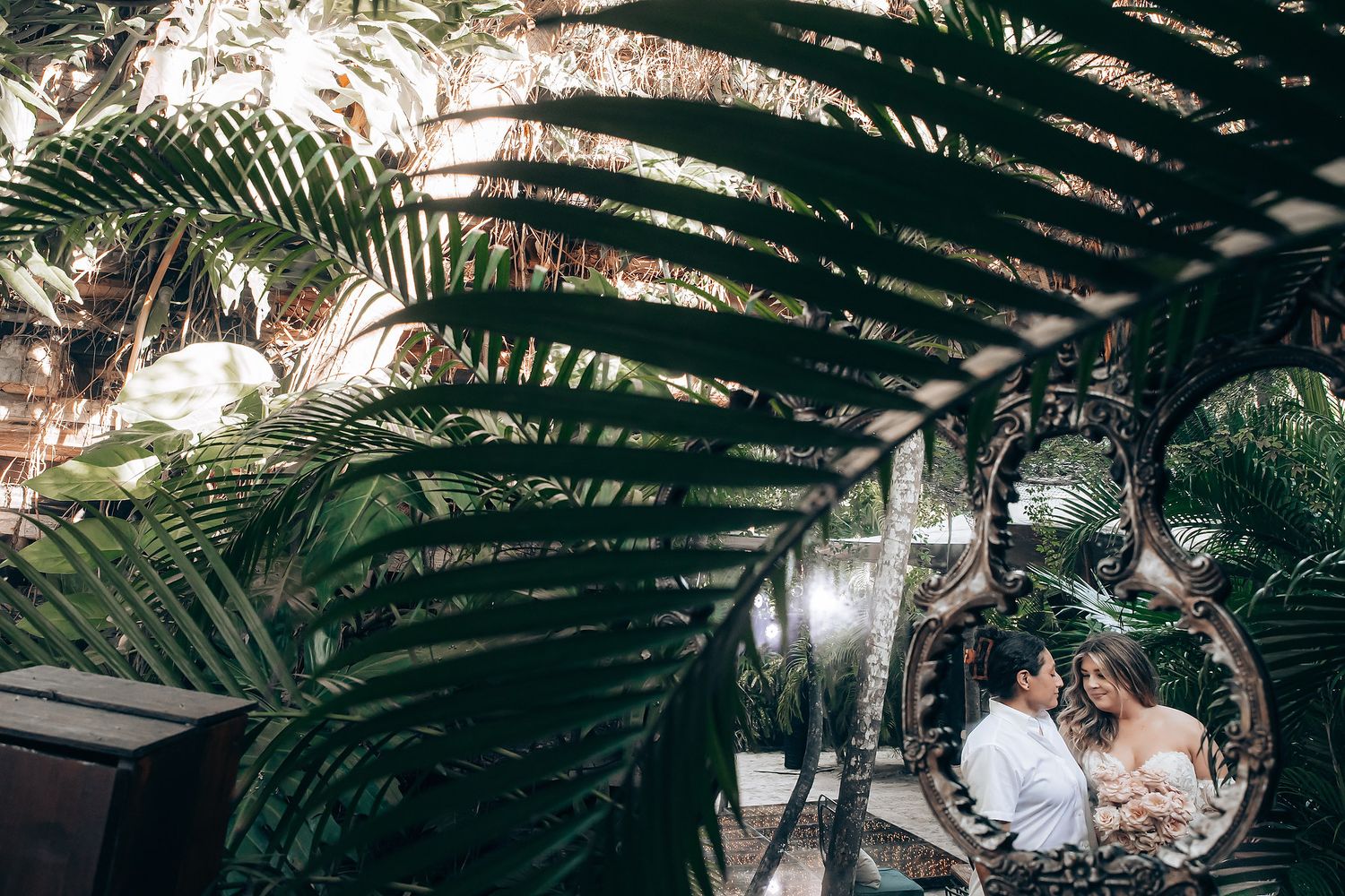 A romantic wedding couple shares an intimate moment under tropical palm fronds at a garden venue.