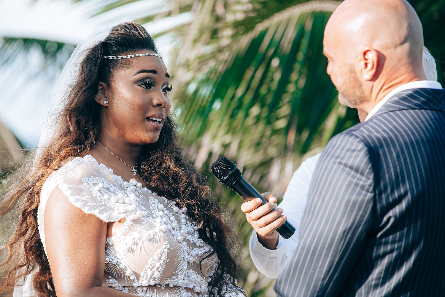 A bride in a lace wedding dress stands with an officiant holding a microphone during an outdoor tropical ceremony.