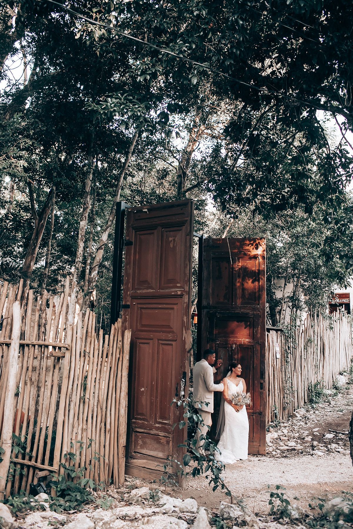 A rustic wedding venue entrance features wooden doors and bamboo fencing surrounded by lush greenery.