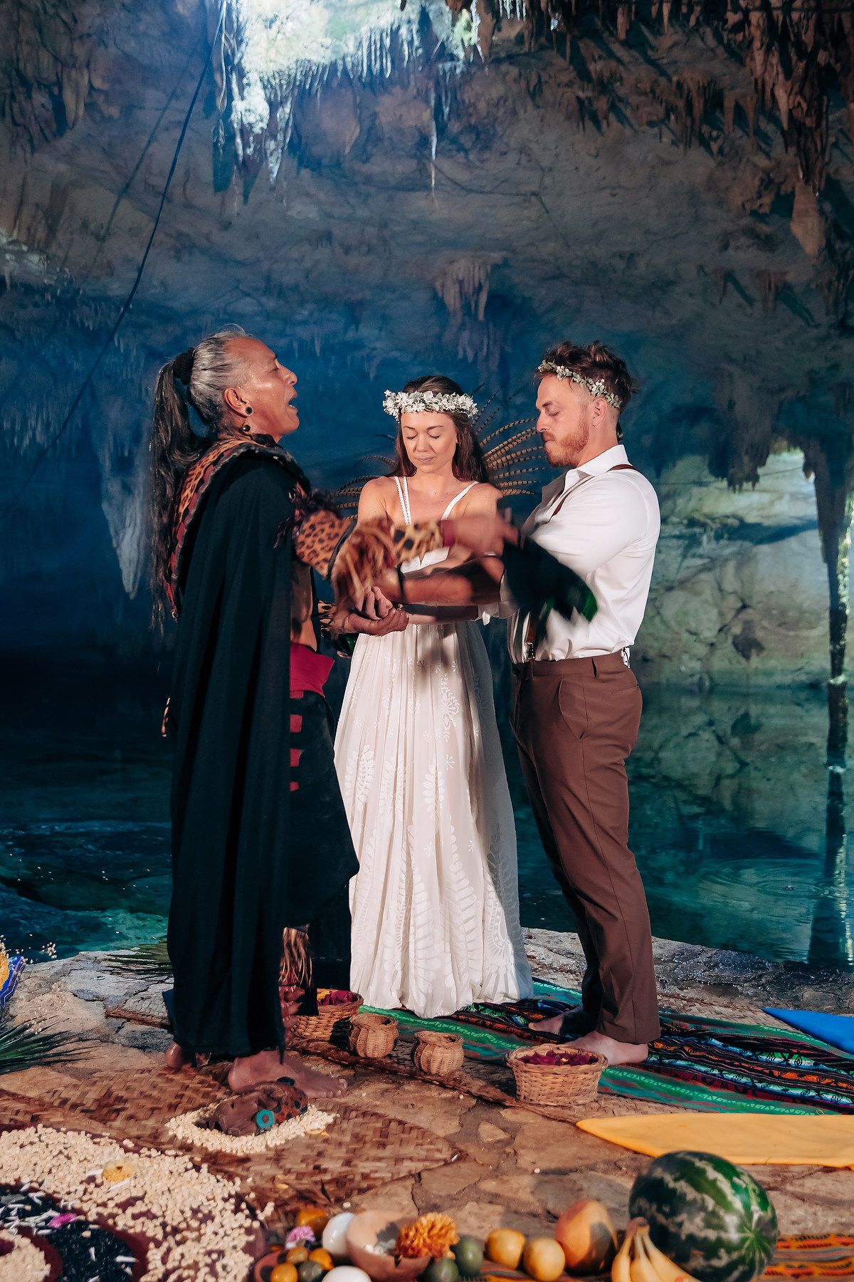 A wedding ceremony takes place in a dramatic cave setting with colorful textiles and decorations on the stone floor.