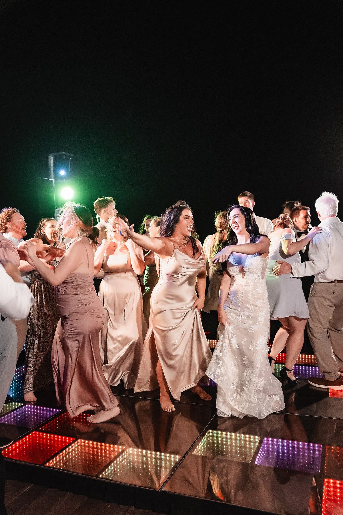 Wedding party dances together on illuminated dance floor during evening reception celebration.