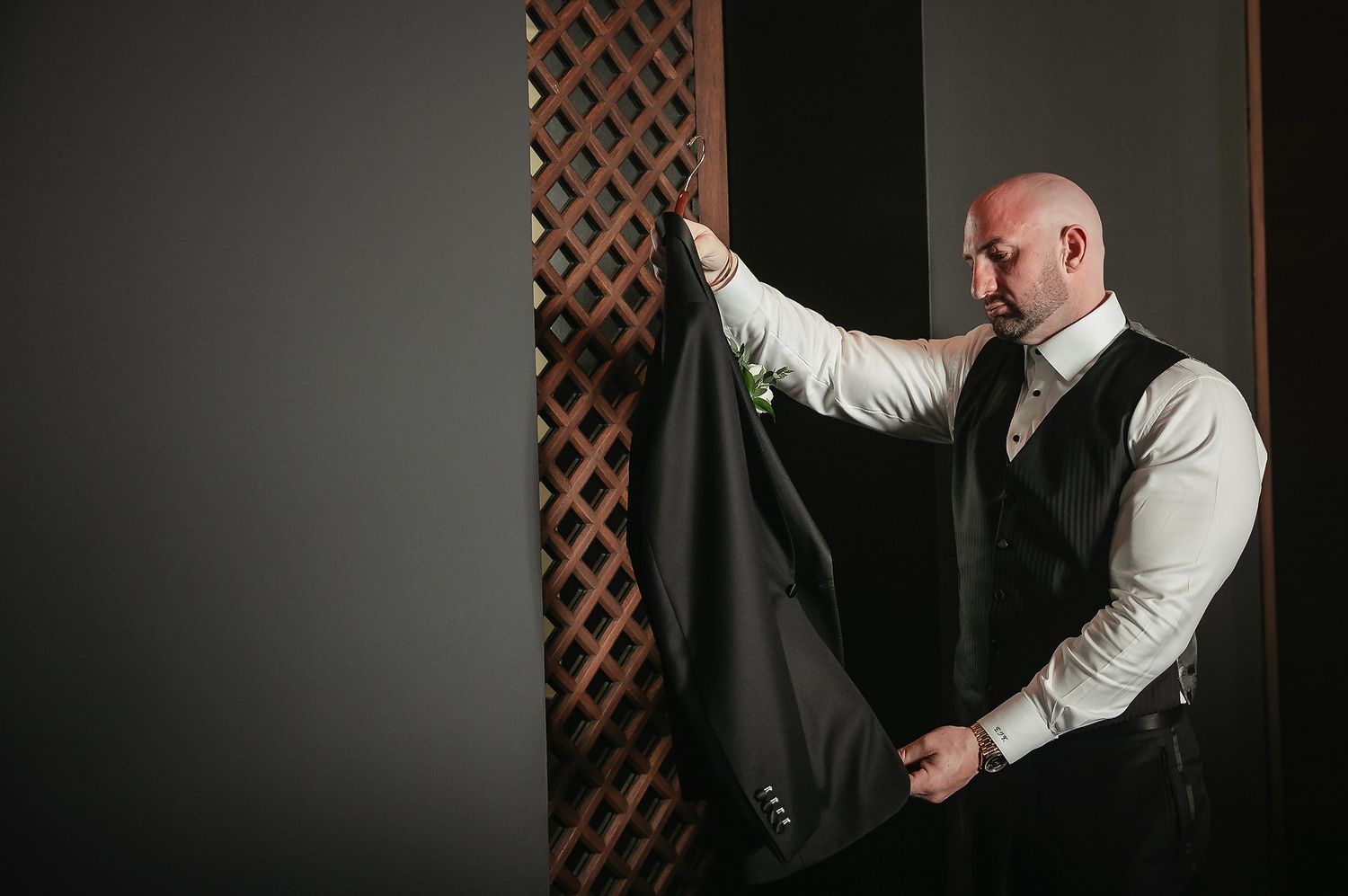 A person in a white dress shirt and black vest adjusts a dark garment while standing near a lattice wall.