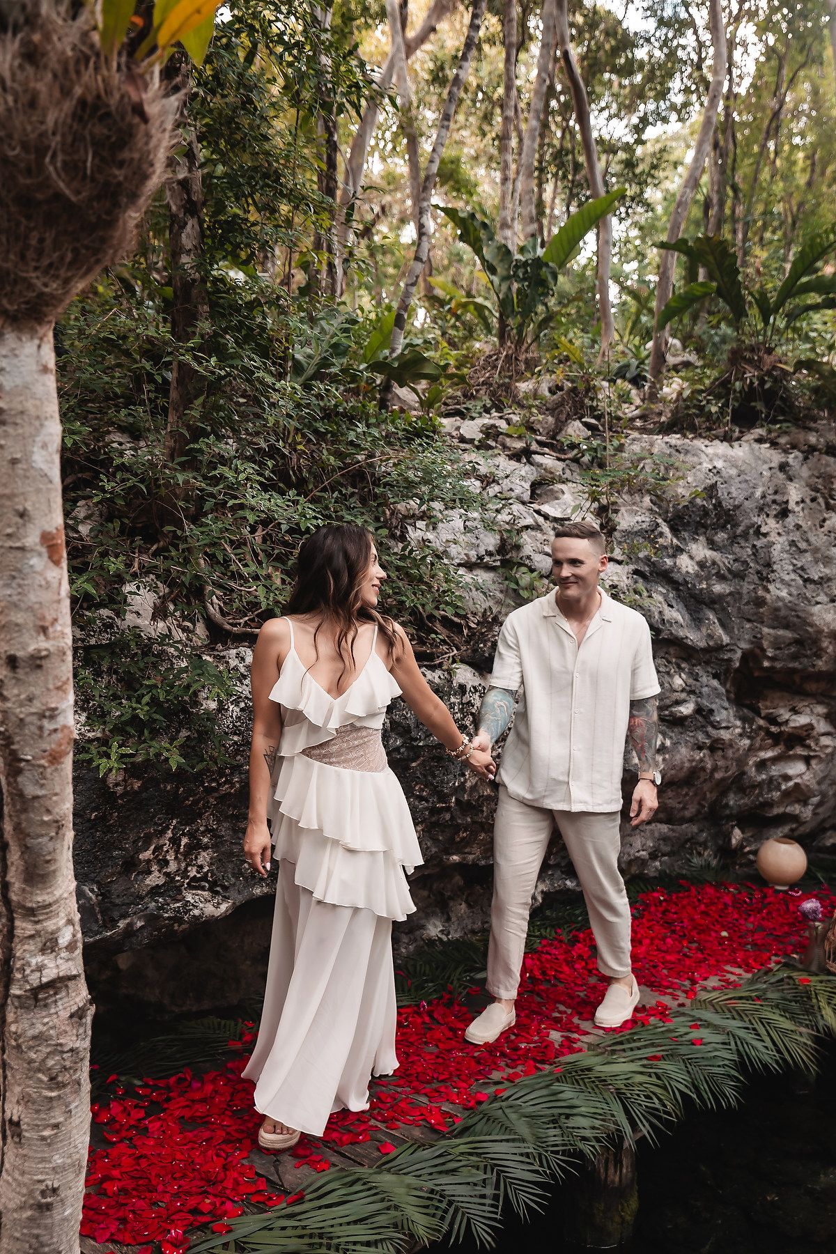 Romantic outdoor scene with white outfits against stone walls and scattered red rose petals.