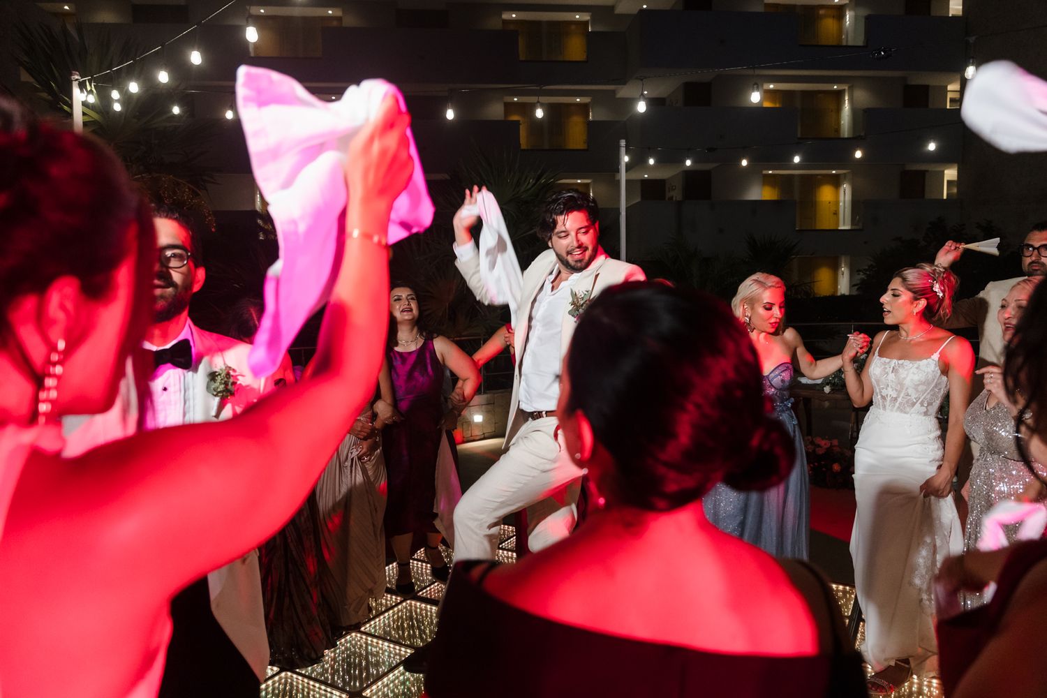 Groom in white suit dancing joyfully with wedding guests at a vibrant outdoor reception party at night.