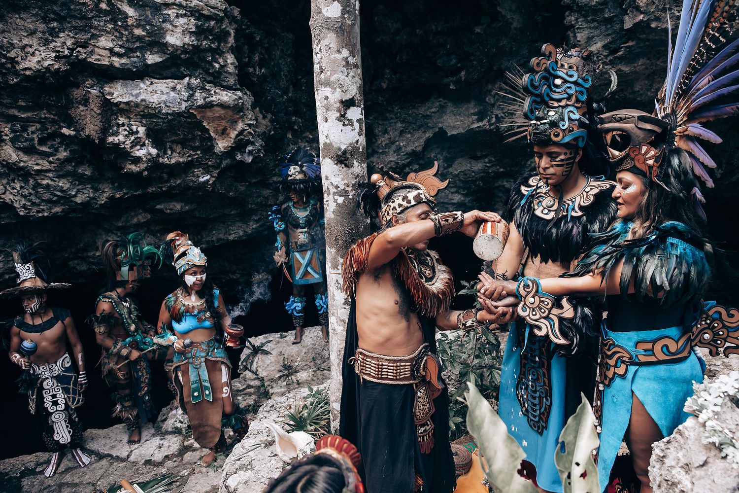 Group of Mayan ceremonial dancers in vibrant turquoise and black feathered costumes prepare near a rocky cave wall.