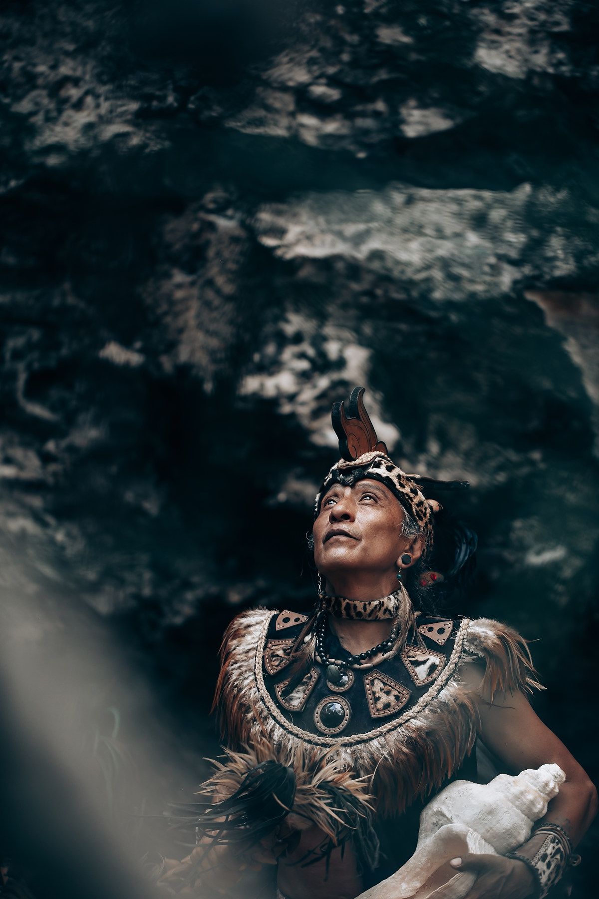 Man in traditional Mayan feathered costume holding conch shell gazing upward near rocky cave wall.