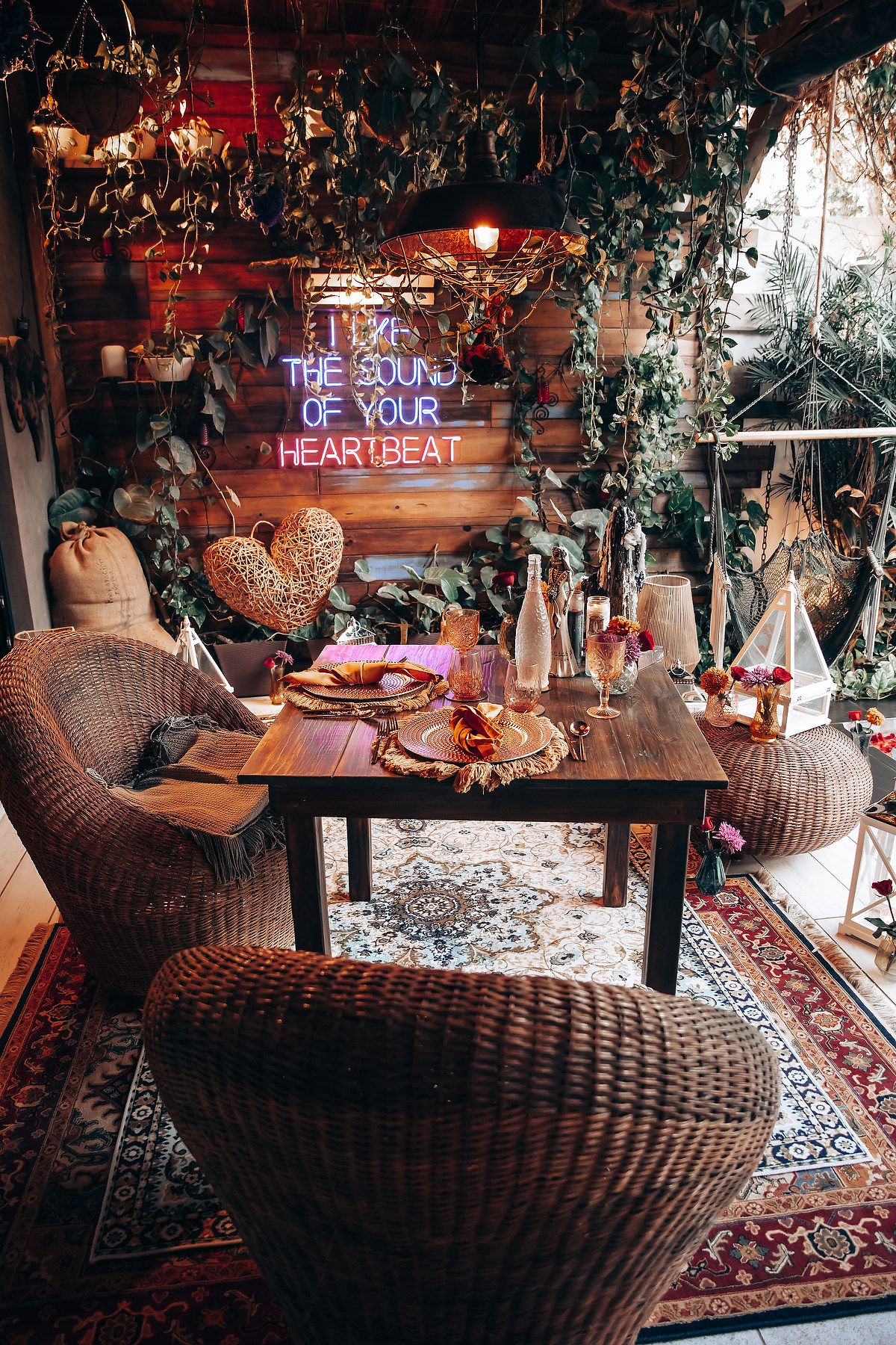 Bohemian restaurant patio with wicker chairs, wooden table, layered rugs, and neon sign reading I Like The Sound Of Your Heartbeat.