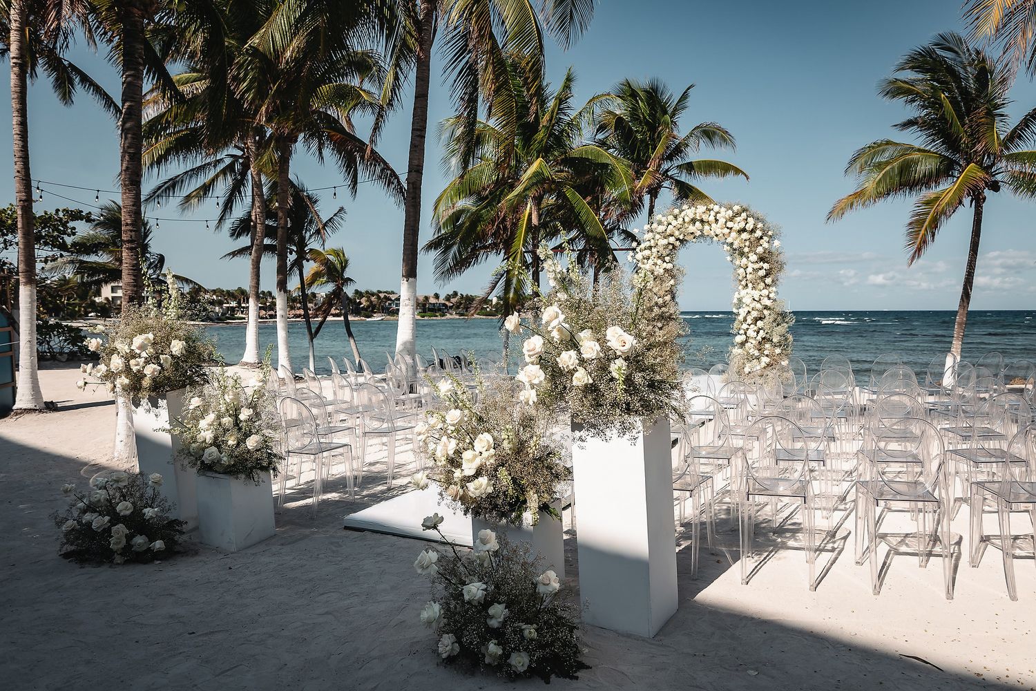 Elegant beachfront wedding ceremony setup with white floral arch, ghost chairs, and palm trees under a clear blue sky.