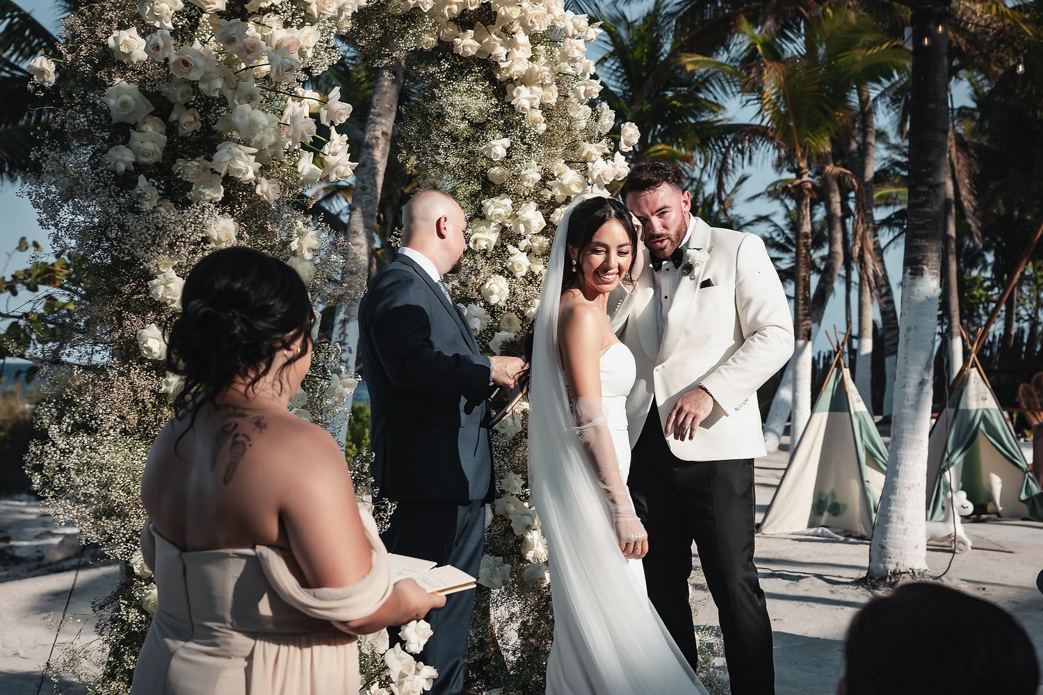 Couple exchanges vows at outdoor beach wedding ceremony beneath white floral arch with palm trees in background.