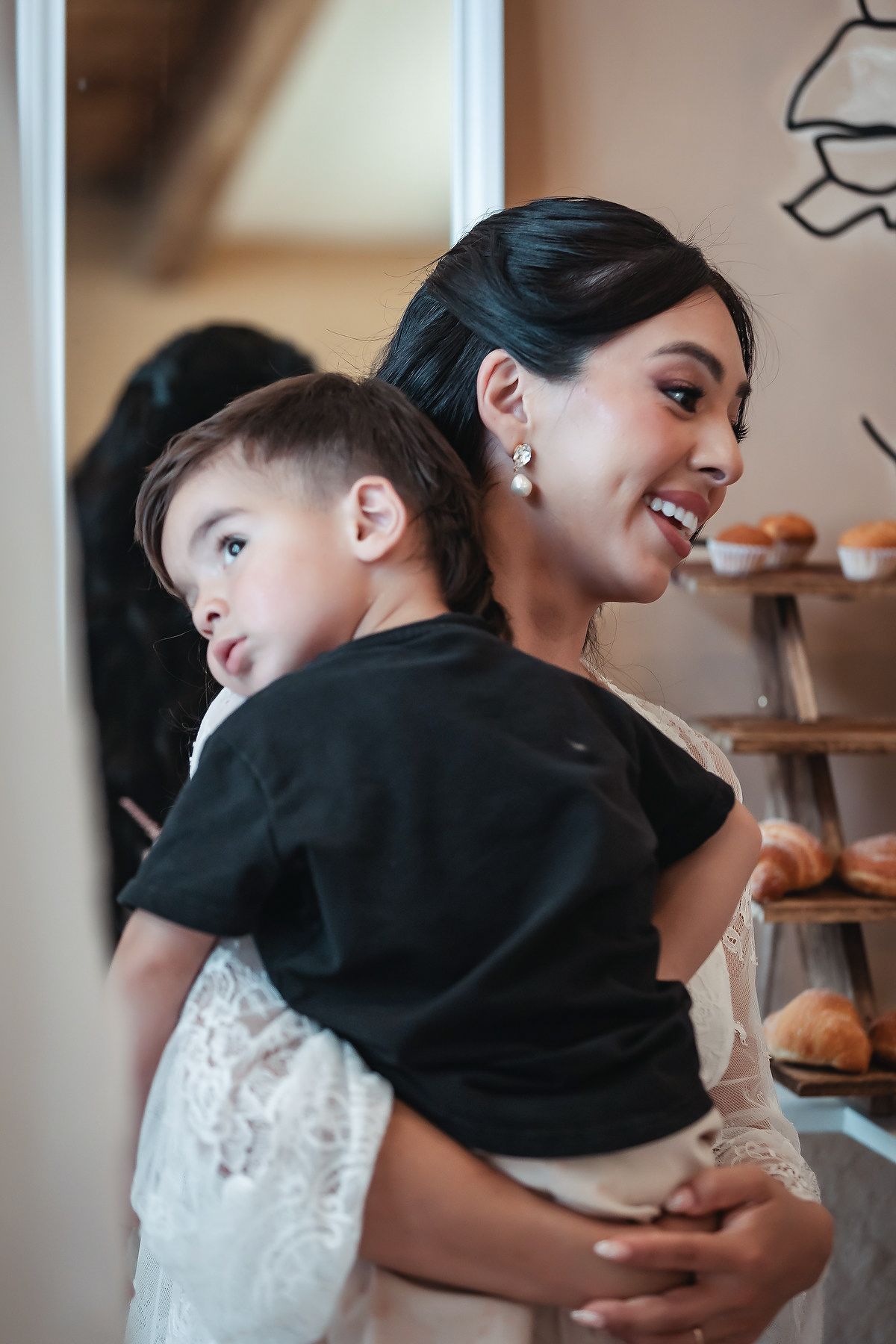 Smiling bride in white lace dress holding a young boy near a rustic bakery display of croissants.