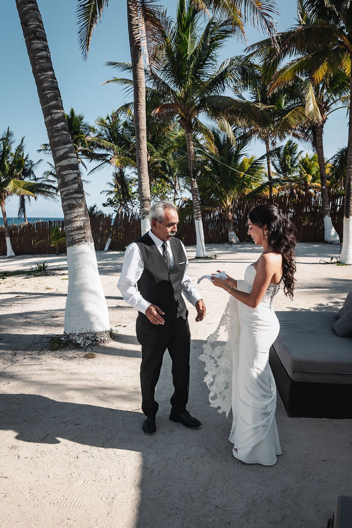 Bride in white gown exchanges vows with groom on tropical beach surrounded by palm trees.