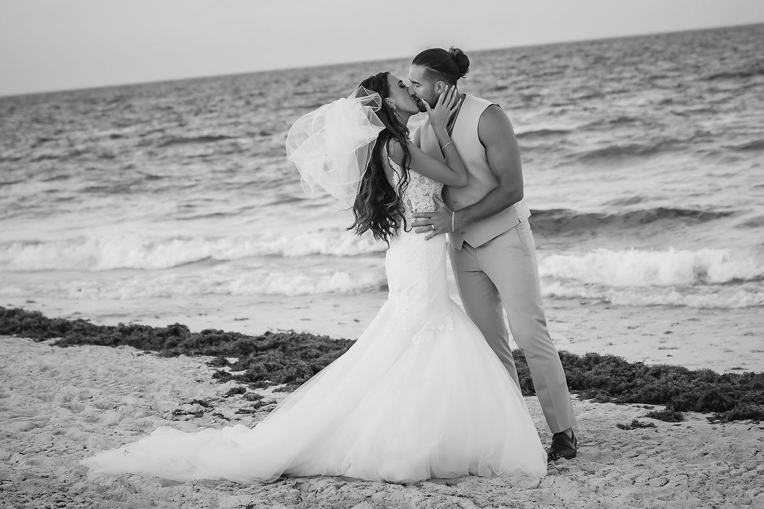 Black and white photo of a bride and groom sharing a romantic kiss on a beach with ocean waves behind them.