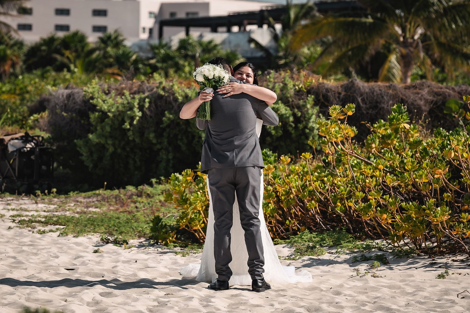 Bride and groom share a romantic beach hug, with lush greenery and a resort in the background.