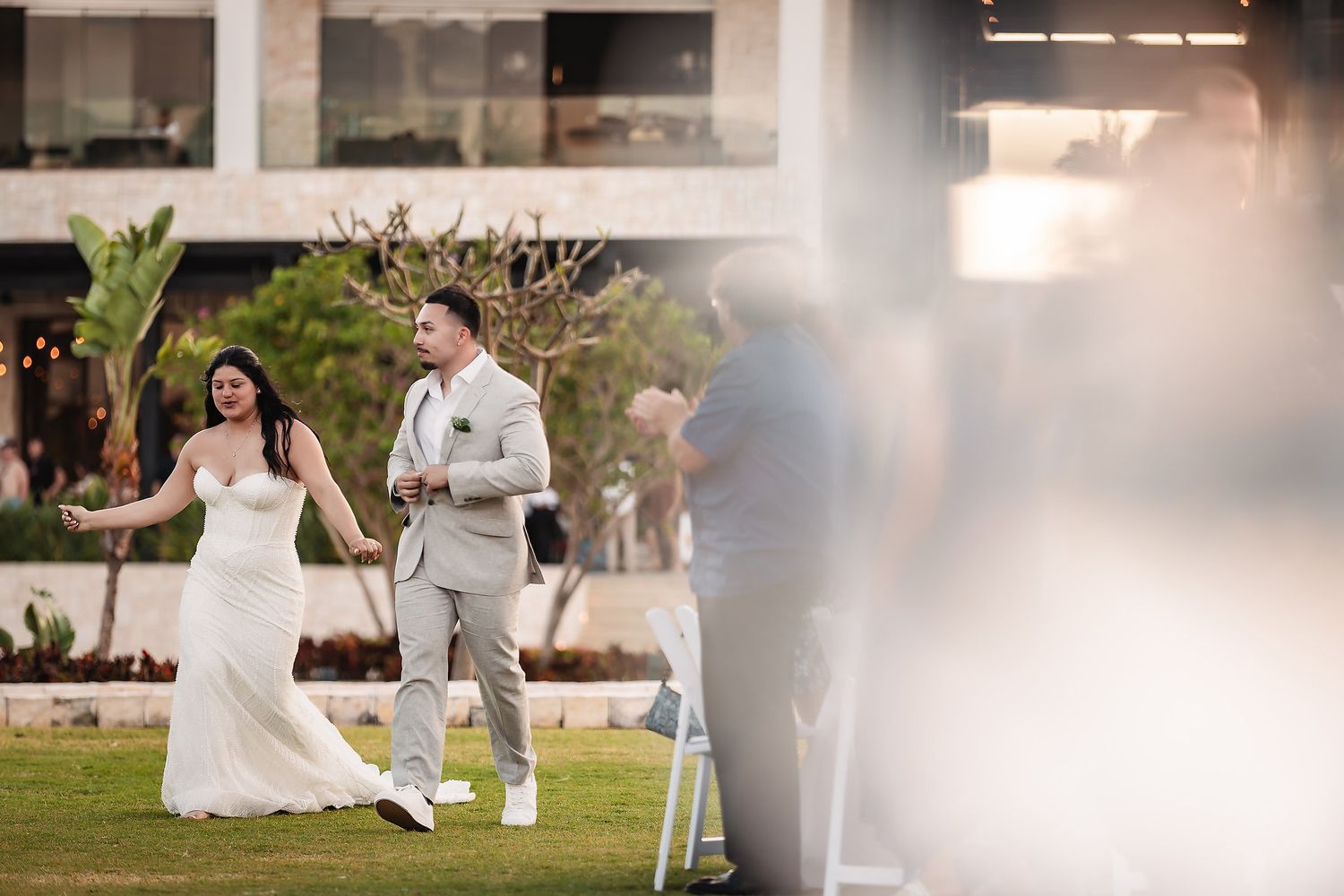 Bride and groom walk hand-in-hand laughing during outdoor wedding reception on tropical resort lawn.