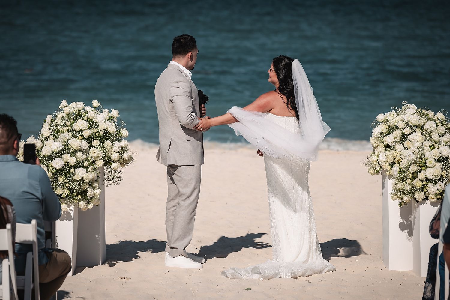 Couple exchanges vows at a romantic beach wedding ceremony with white floral arrangements and ocean backdrop.
