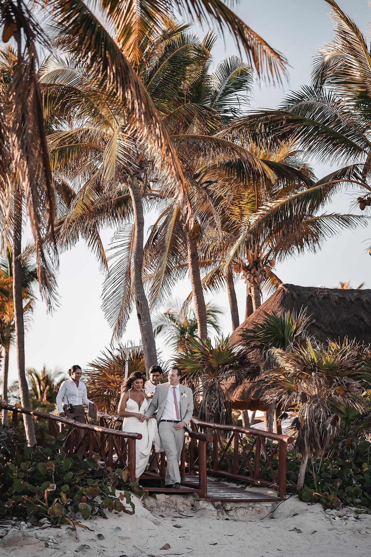 Bride and groom walk on a wooden beach path surrounded by tall palm trees at a tropical destination wedding.