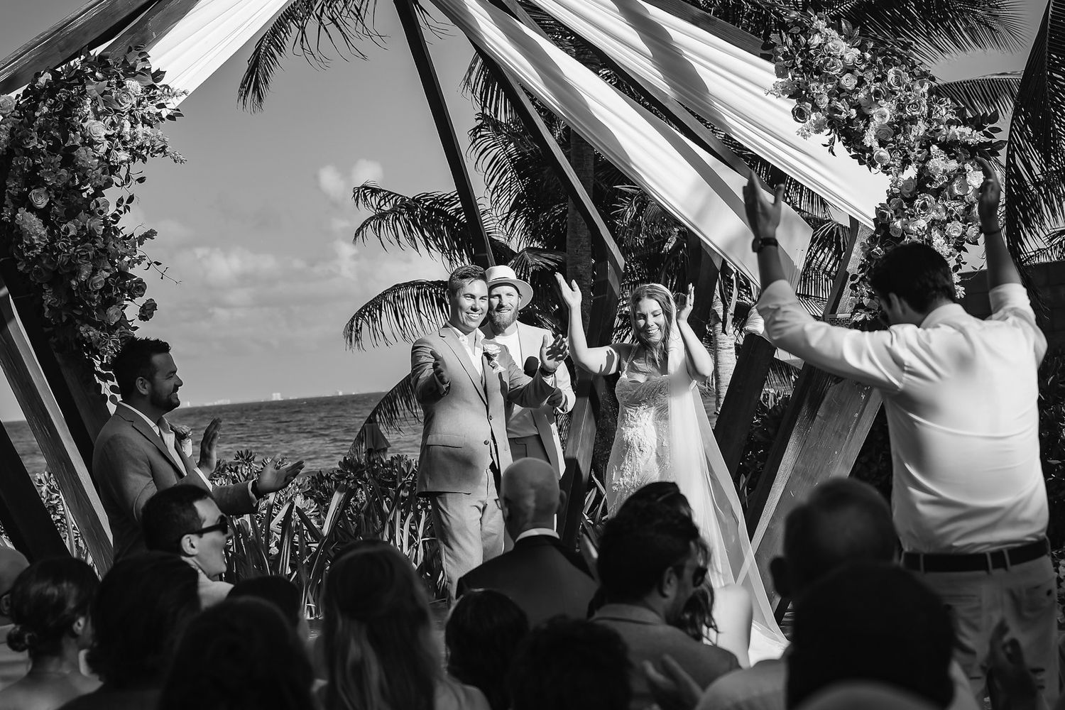 Black and white photo of a joyful wedding ceremony outdoors under a fabric canopy with palm trees in the background.