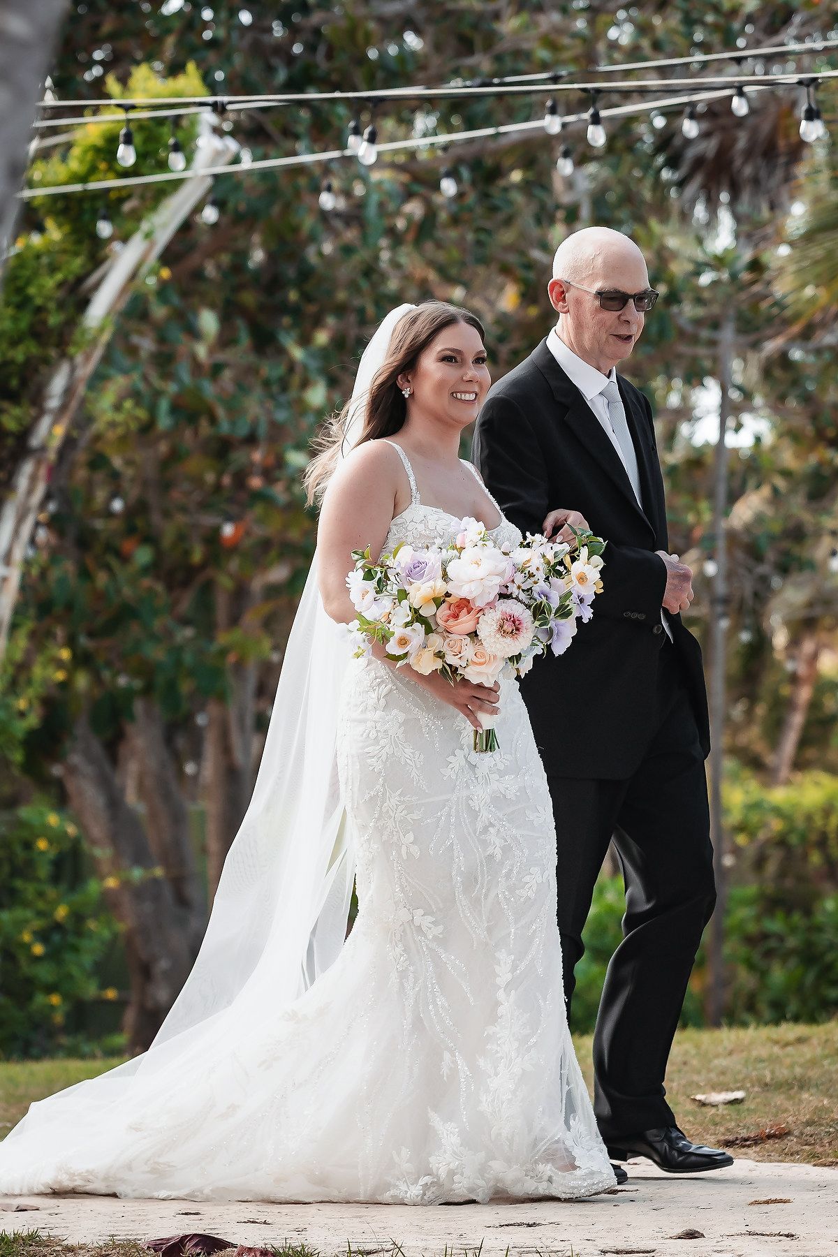 Bride in white lace gown holding pastel bouquet walks down aisle with bald man in black tuxedo at outdoor wedding.