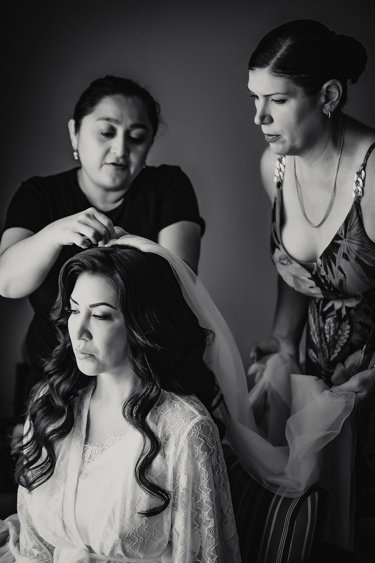 Black and white photo of a bride getting her veil placed by two women during wedding preparations.