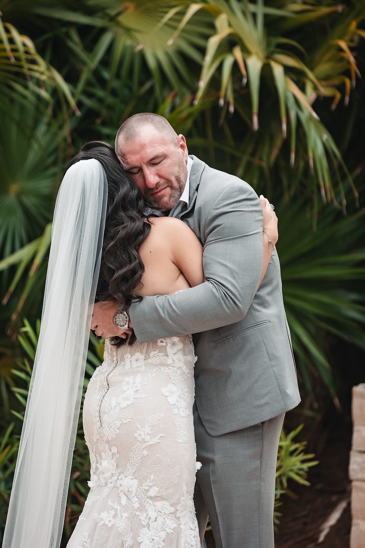Emotional groom in gray suit embracing bride in lace wedding dress with veil, sharing tender first look moment outdoors.