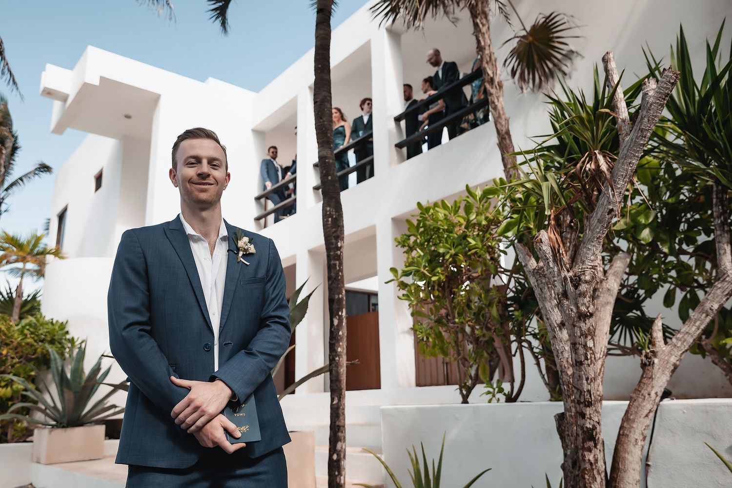 Groom in blue suit standing outdoors at a white resort with groomsmen on balcony.