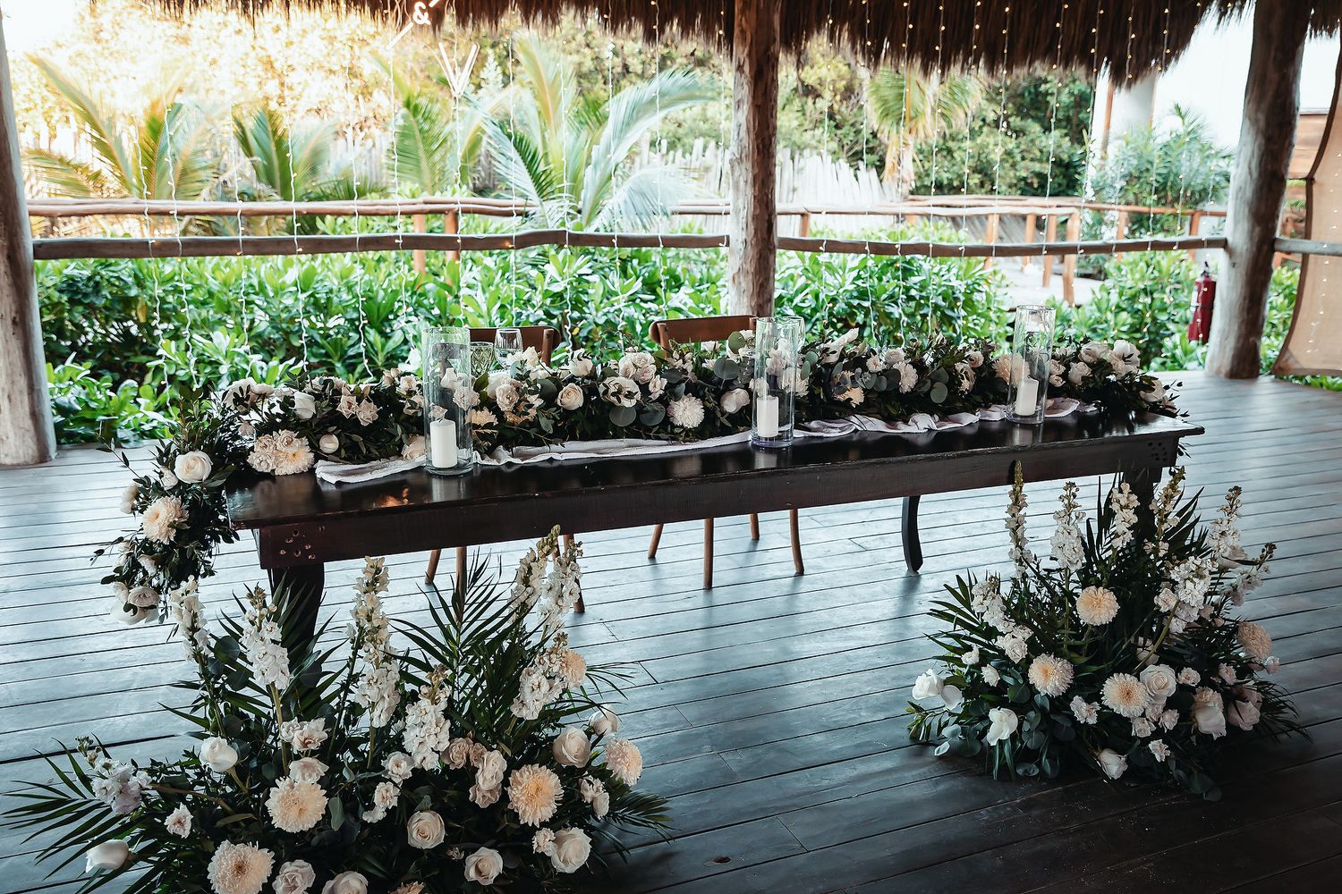 Elegant outdoor wedding head table adorned with white floral arrangements and tropical greenery under a thatched roof pavilion.