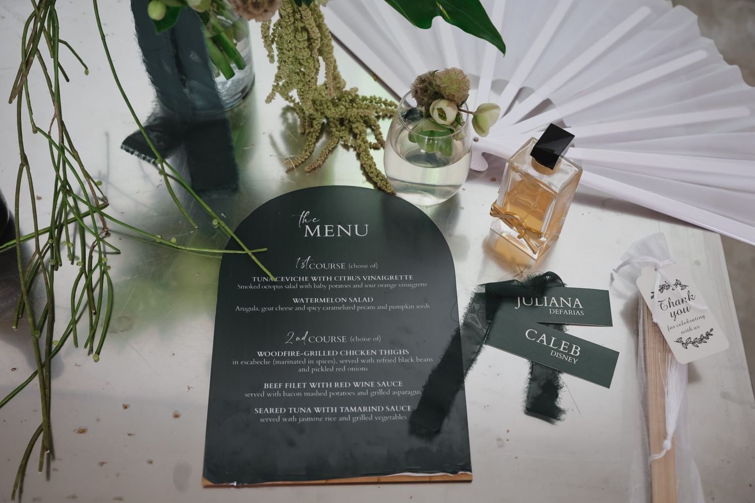 Elegant wedding stationery flat lay featuring a dark green menu card, name cards, and floral accents on a white surface.