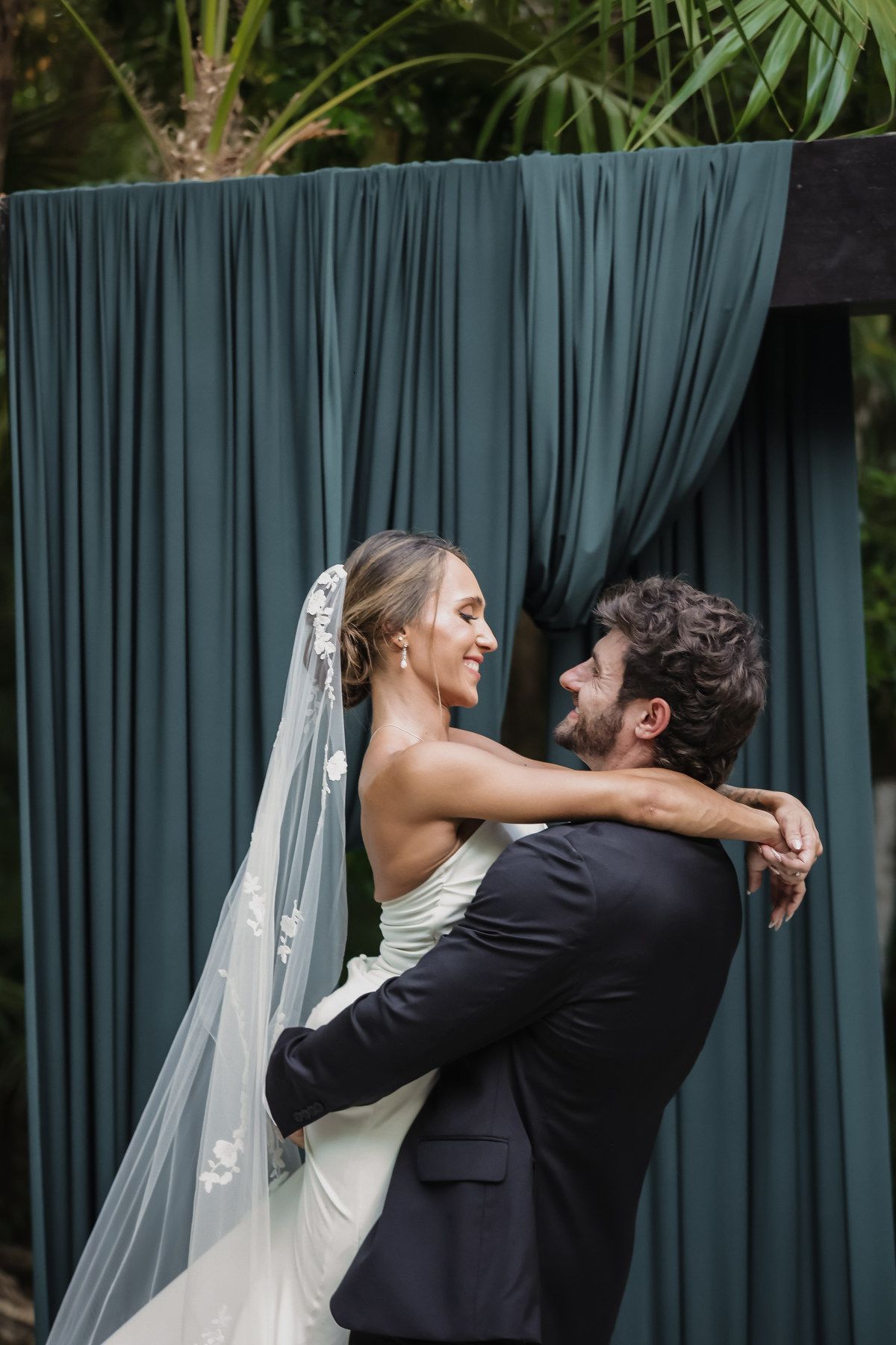 Smiling bride in white gown and veil embraces groom in dark suit before elegant teal draped backdrop outdoors.