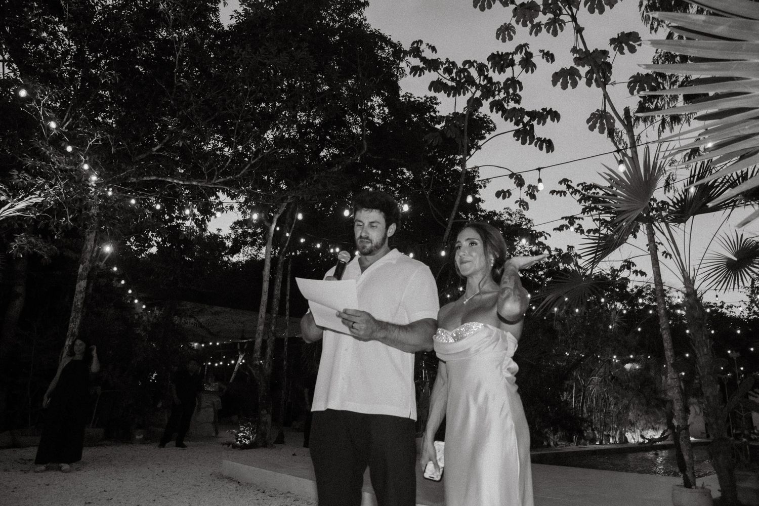 Couple reading wedding vows outdoors at night under string lights surrounded by tropical trees in black and white.