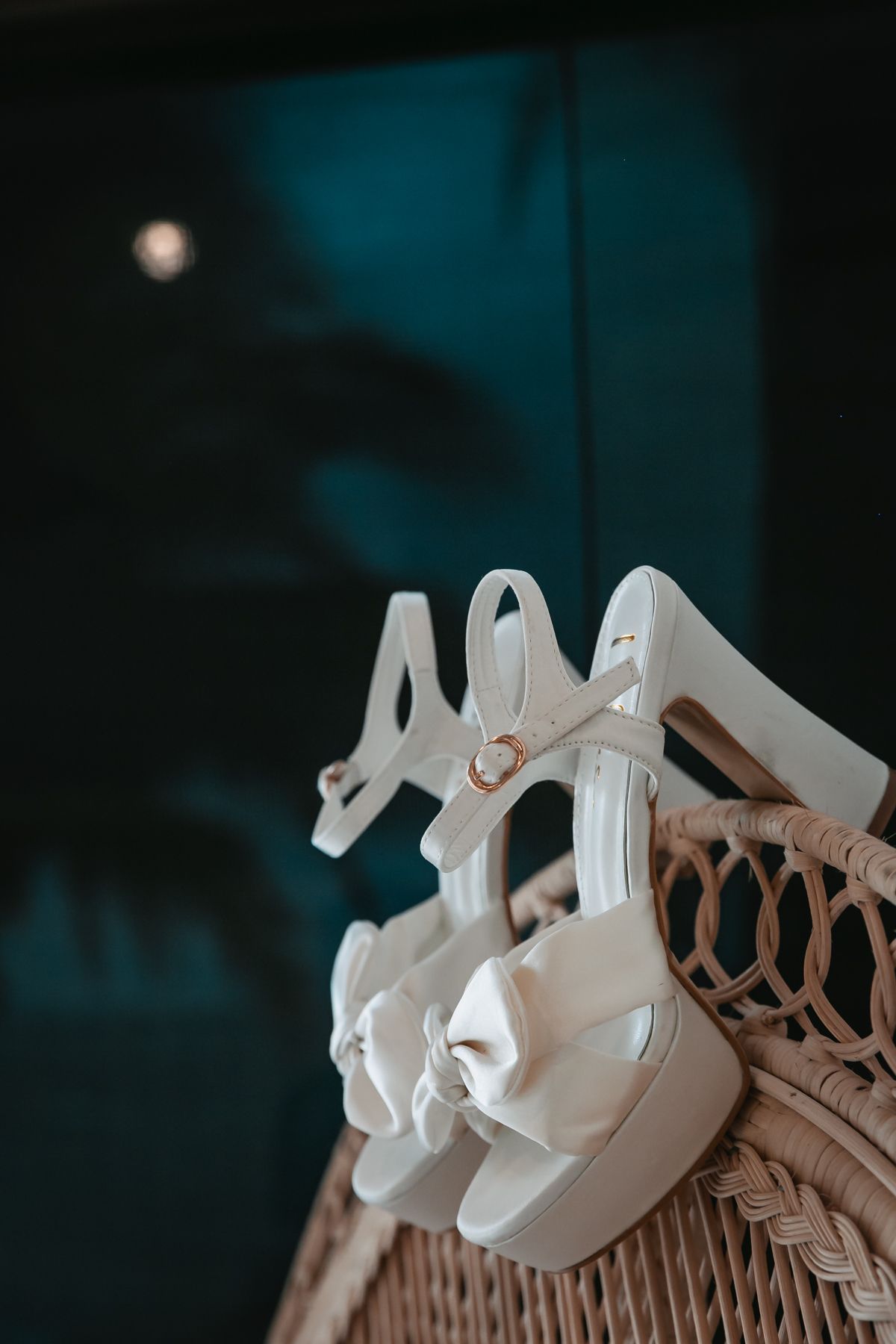 White bridal heels with bow detail and ankle strap displayed on wicker chair at tropical wedding venue.
