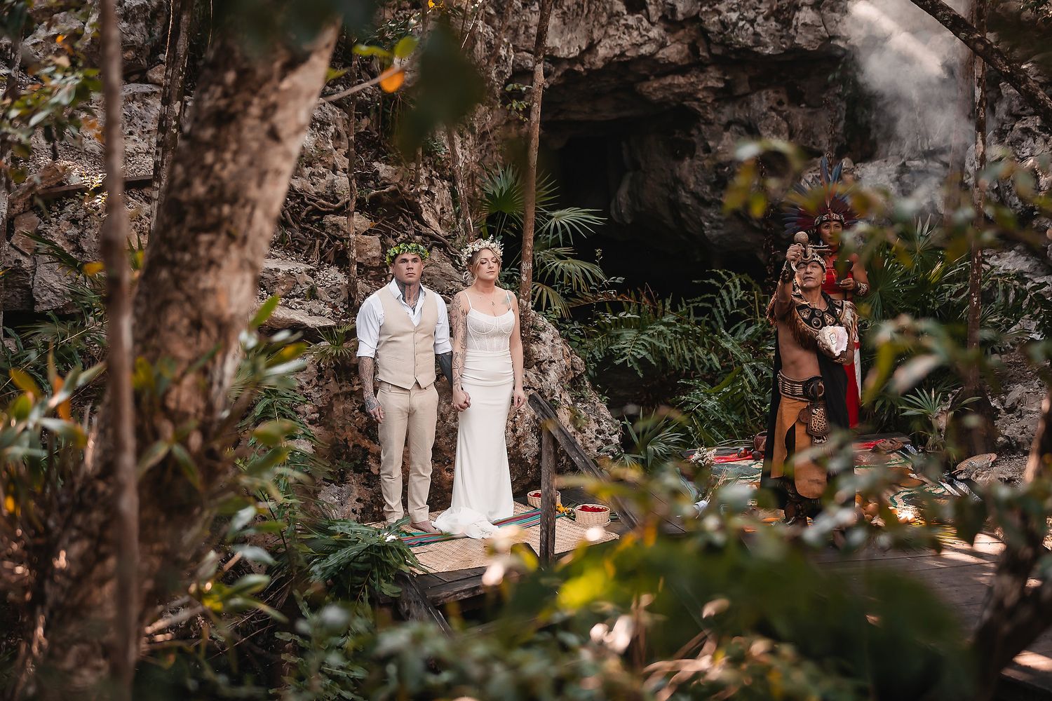 Couple exchanges vows in lush jungle setting with officiant in traditional attire near rocky cave backdrop.