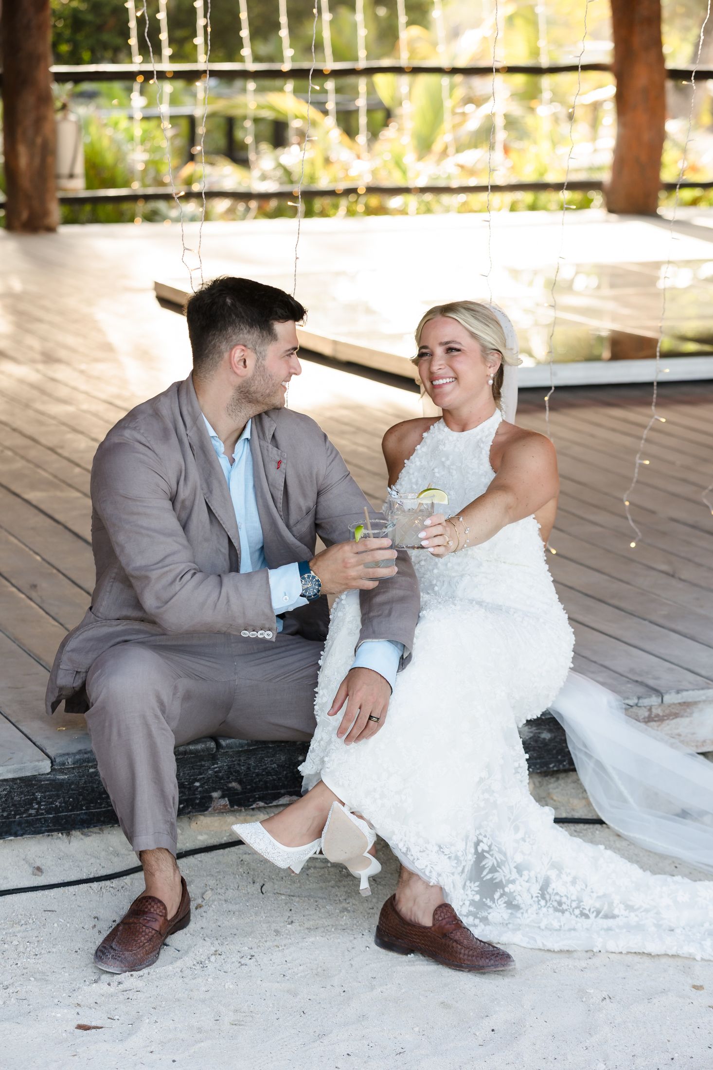 Bride and groom sitting together on wooden deck, clinking glasses and smiling at each other on their wedding day.