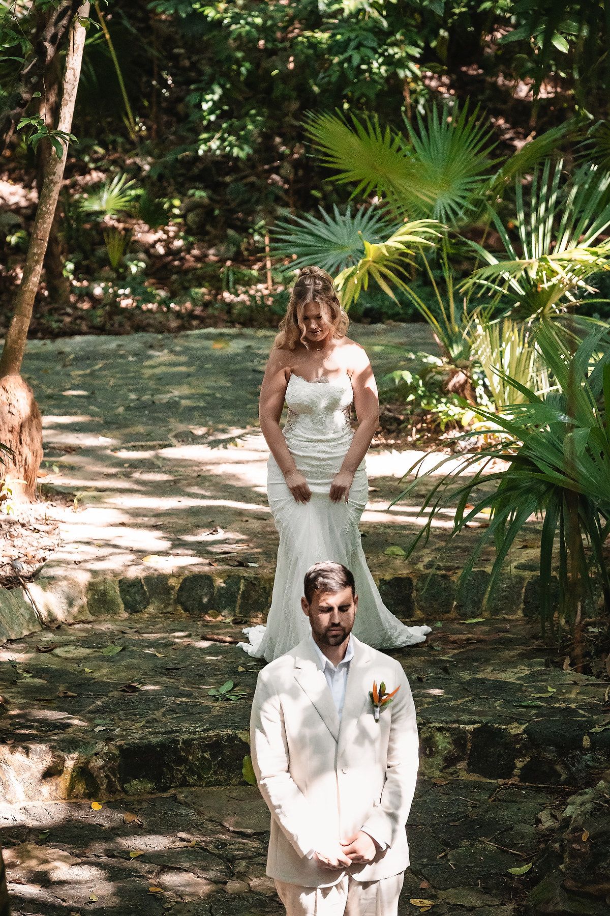 Bride in white gown approaches groom in cream suit during outdoor tropical wedding first look ceremony.