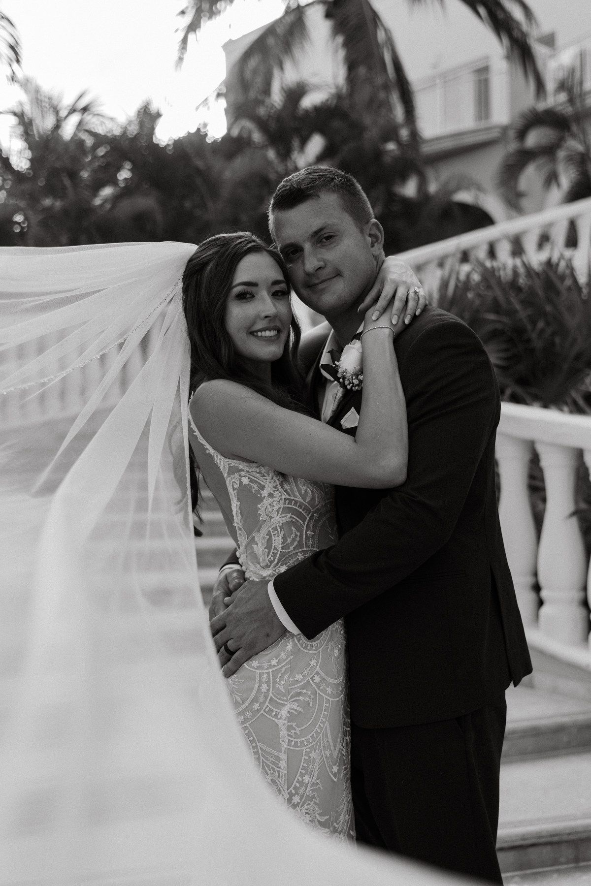 Black and white portrait of bride and groom embracing with flowing veil near white balustrade and palms.
