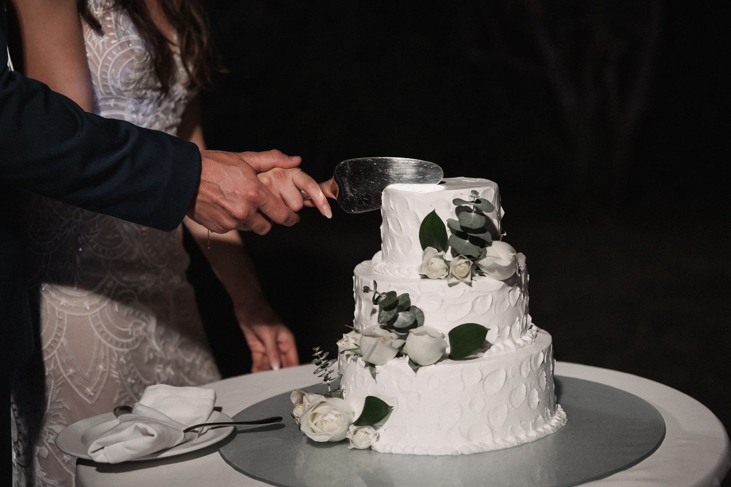 Bride and groom cutting elegant three-tier white wedding cake decorated with white roses and greenery.