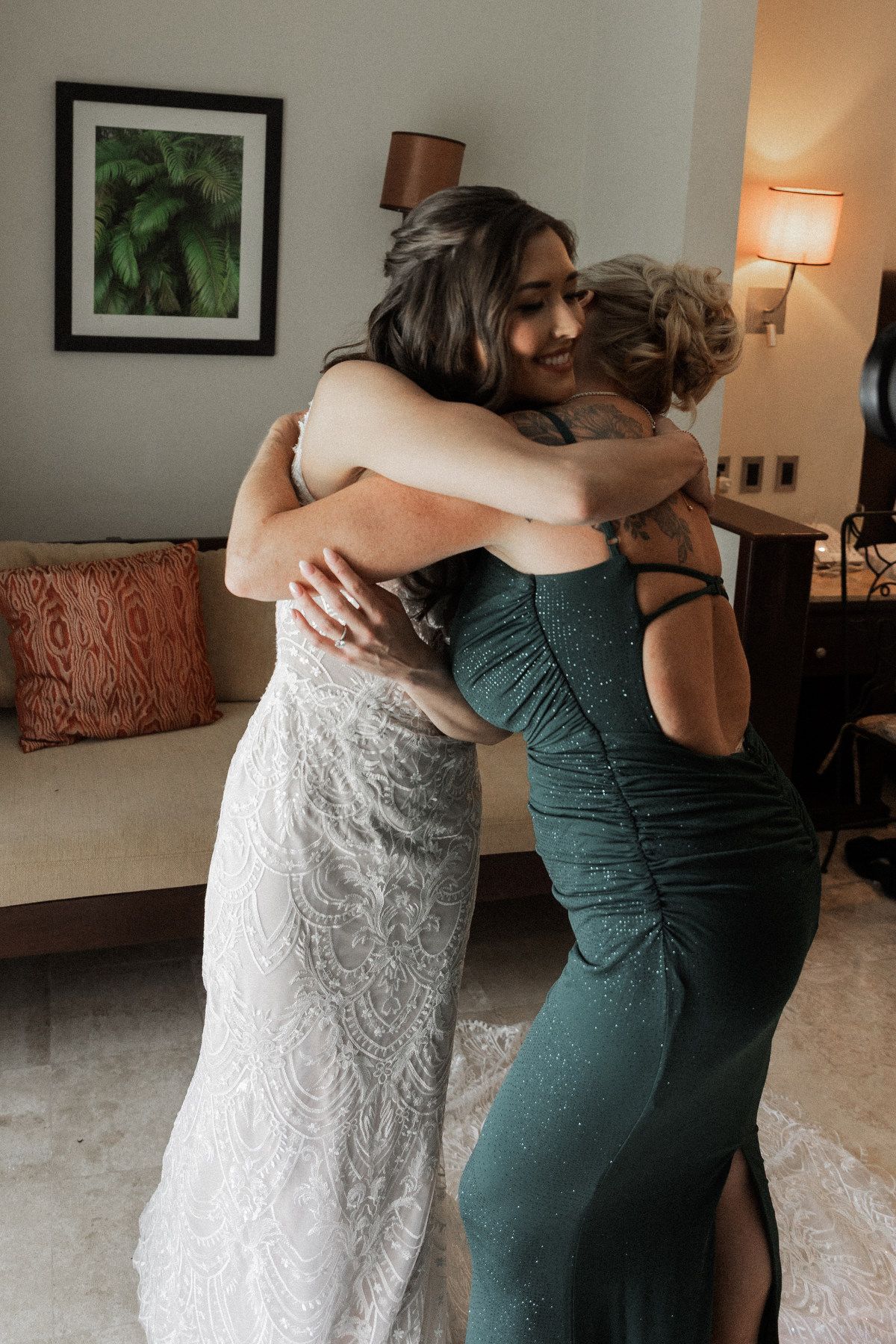 Bride in lace wedding gown sharing an emotional hug with a bridesmaid in a green dress.