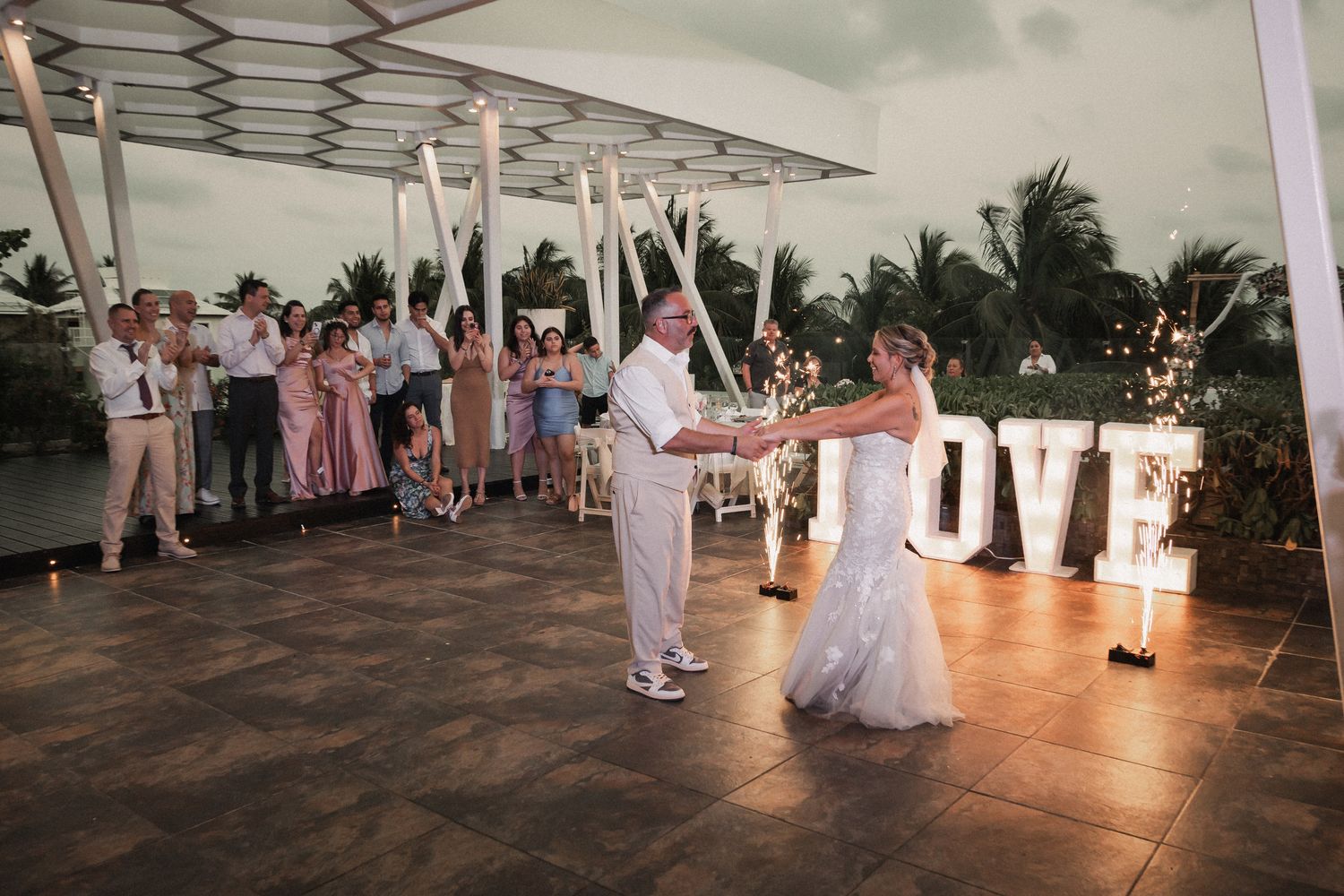 Bride and groom share first dance on outdoor terrace with illuminated LOVE sign at wedding reception.
