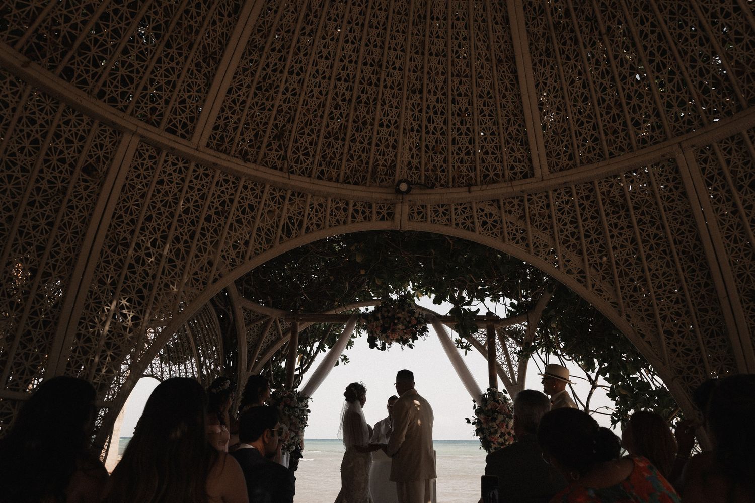 Silhouettes of wedding couple and guests gathered under ornate domed gazebo overlooking tropical beach.