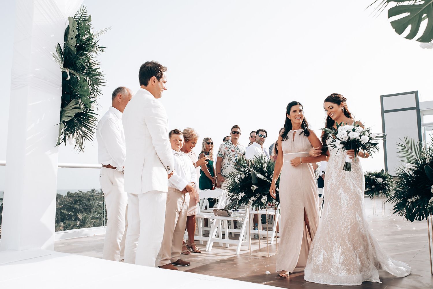 Wedding ceremony setup with white chairs and tropical greenery decorations in an elegant venue.