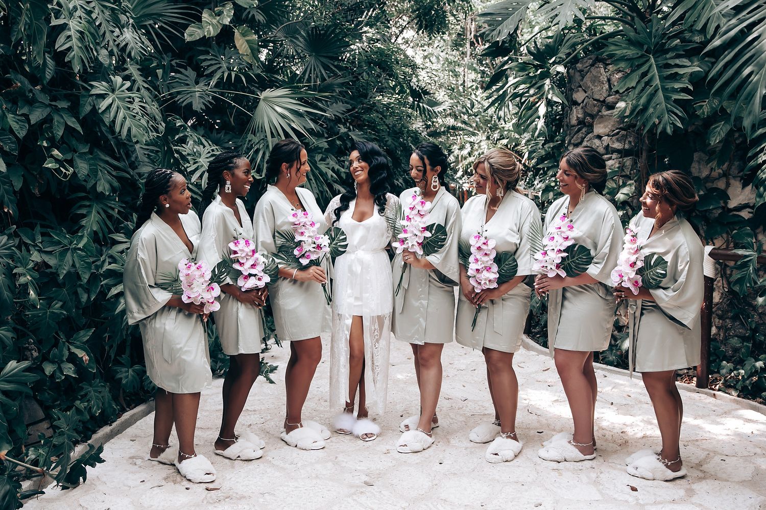 Bridesmaids in white dresses pose in a tropical garden setting with lush green foliage in the background.