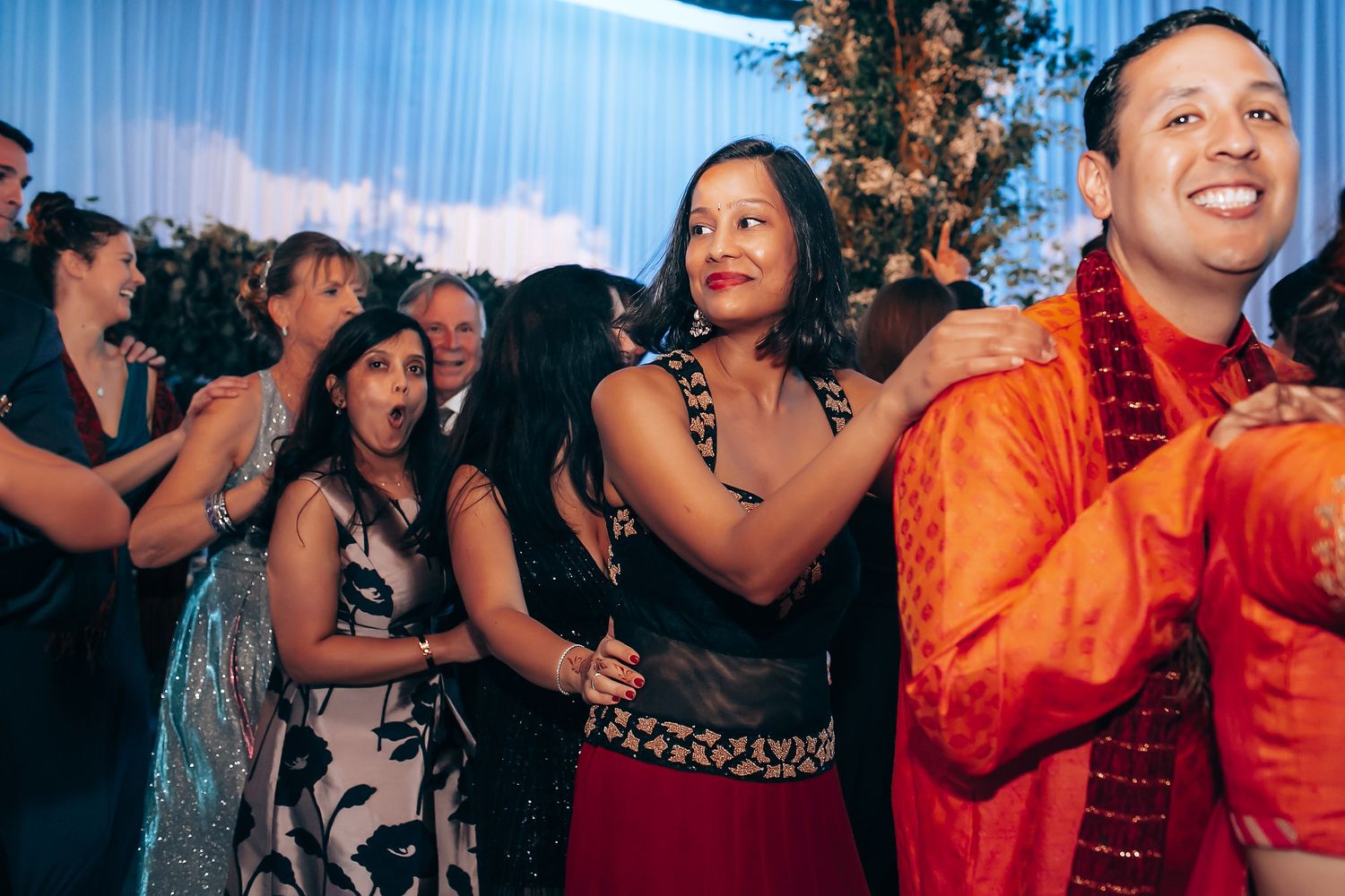 Lively wedding reception with guests dancing under blue lighting and curtained backdrop.
