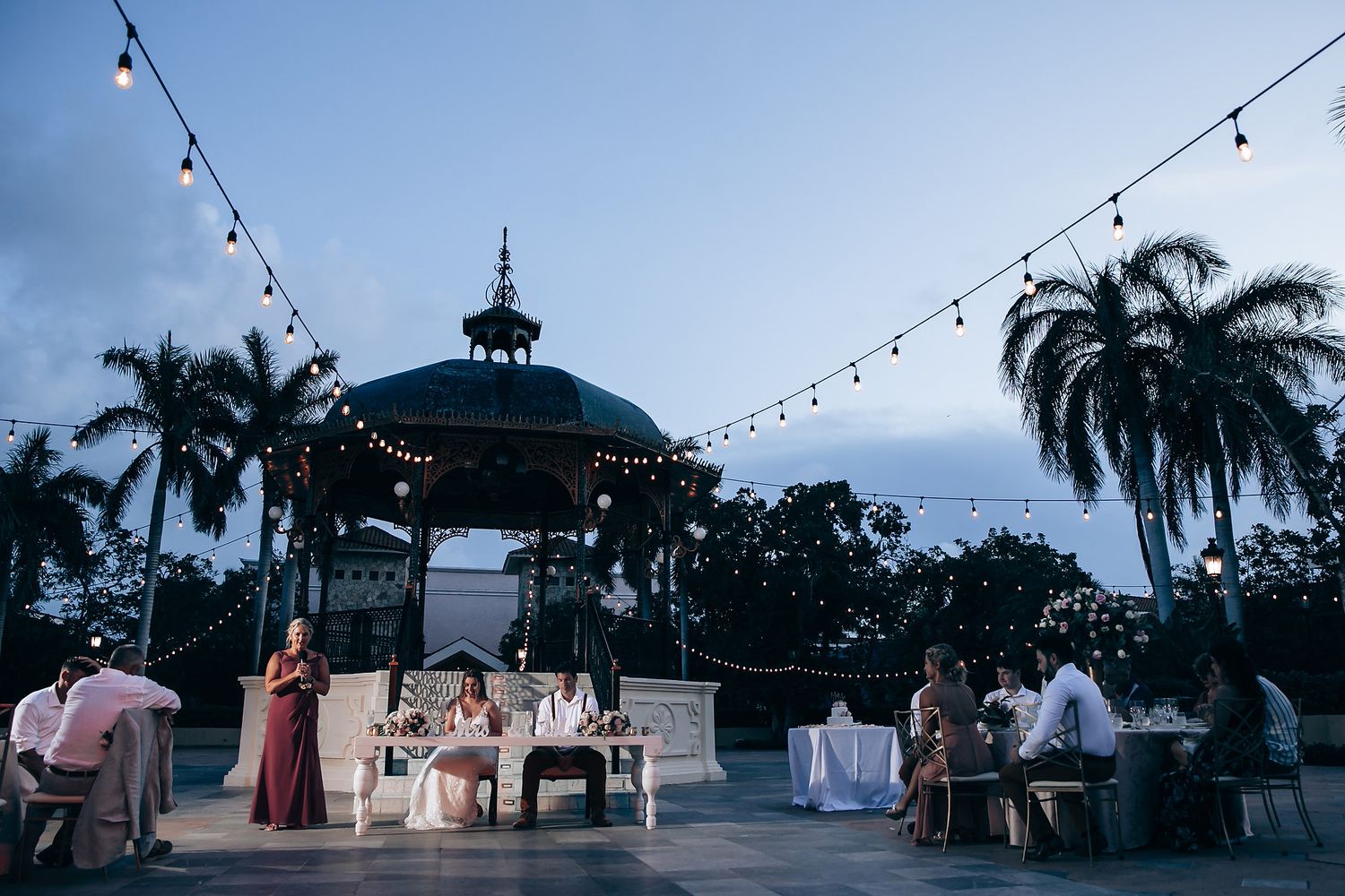 Evening wedding reception setup under a gazebo with string lights and palm trees at dusk.