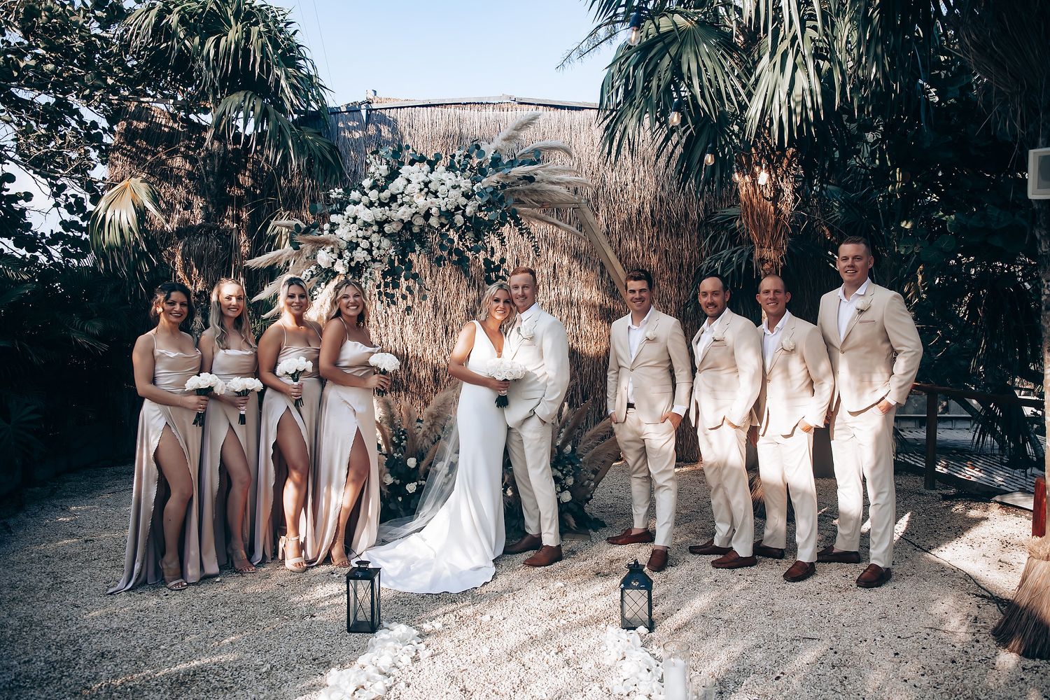 Wedding party lined up outdoors with bridesmaids in beige dresses and groomsmen in light colored suits.