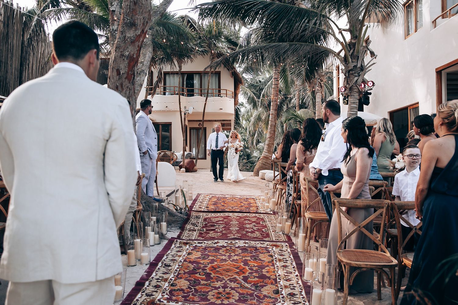 Wedding guests seated outdoors under palm trees with a decorative rug aisle runner.