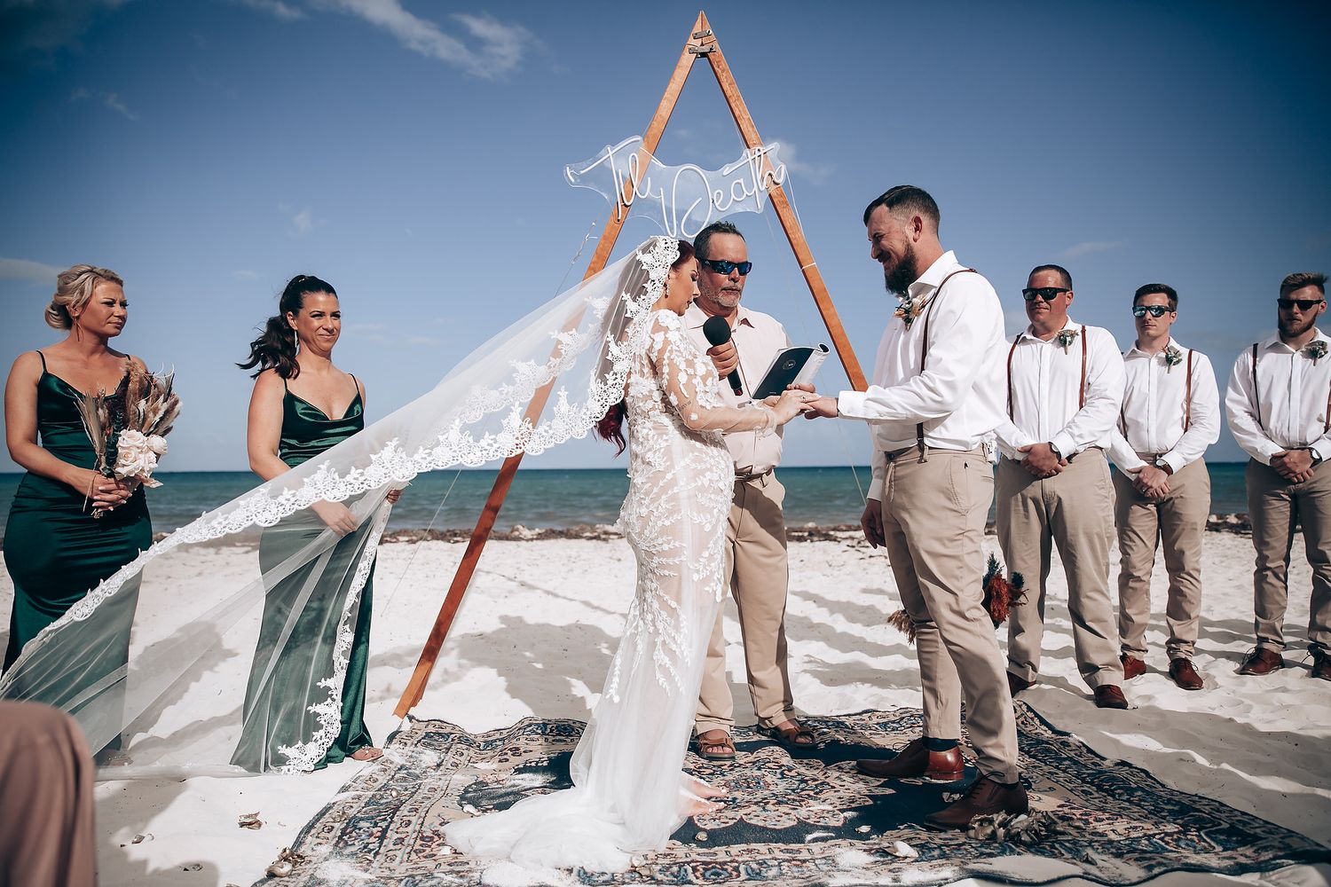 Wedding party gathered on beach with wooden arch and ocean backdrop.