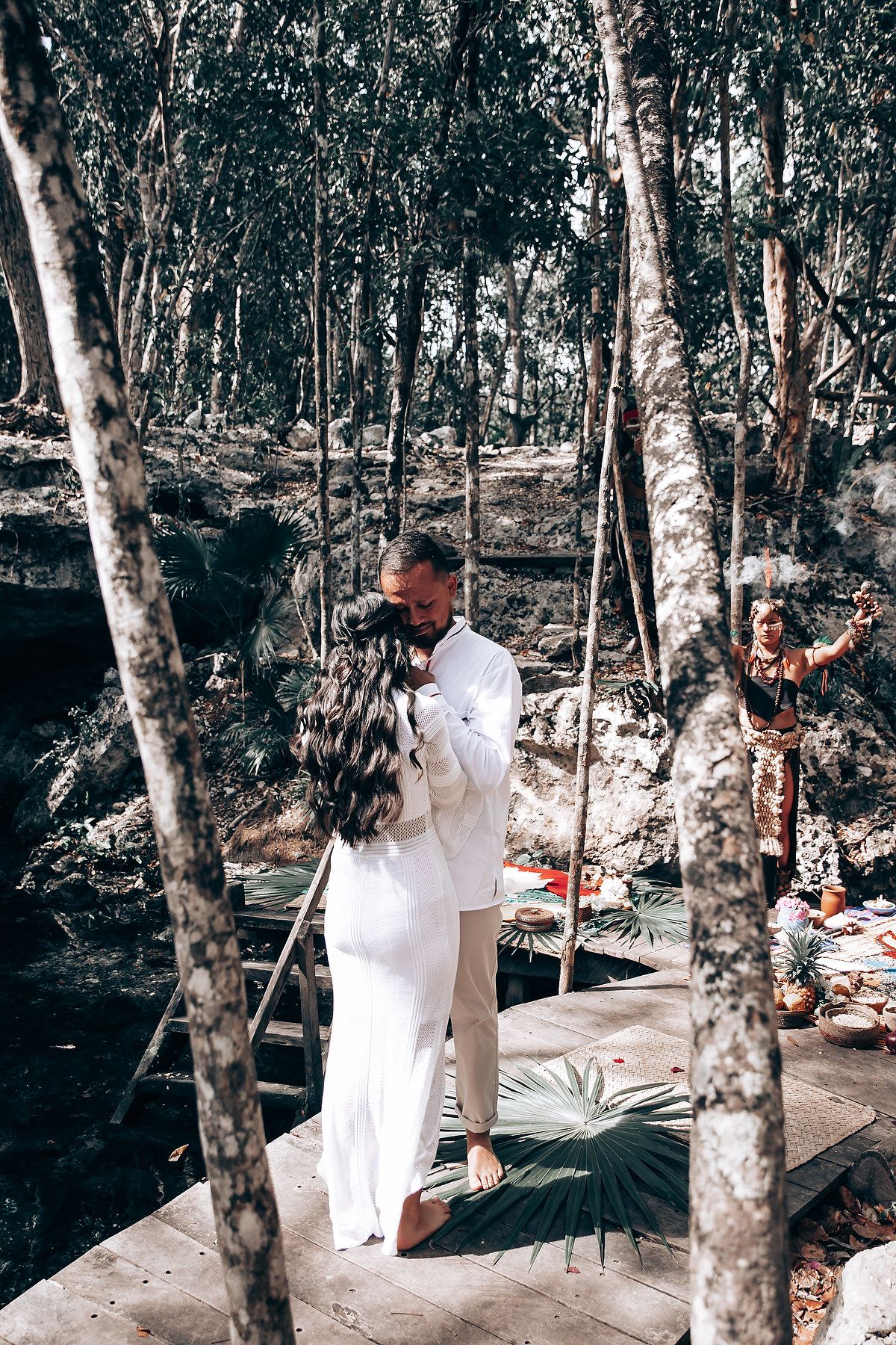 A couple in white attire shares an intimate moment by a serene cenote surrounded by lush forest trees.