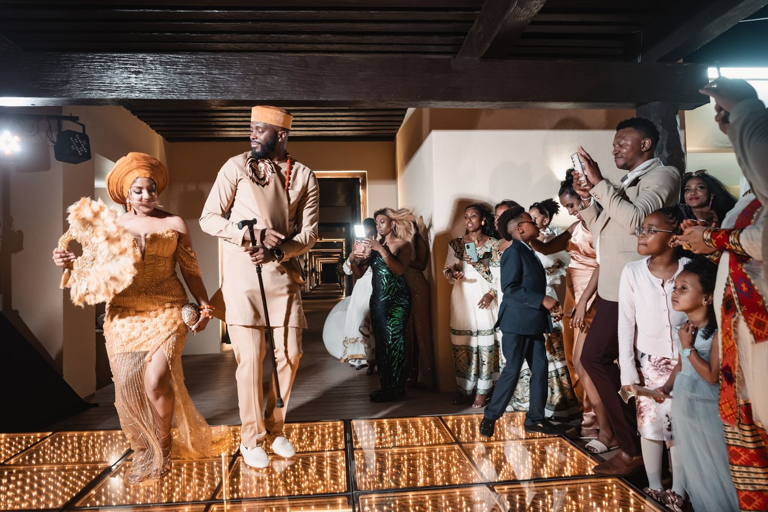 Couple in gold Nigerian traditional wedding outfits dancing on glowing floor with cheering guests.