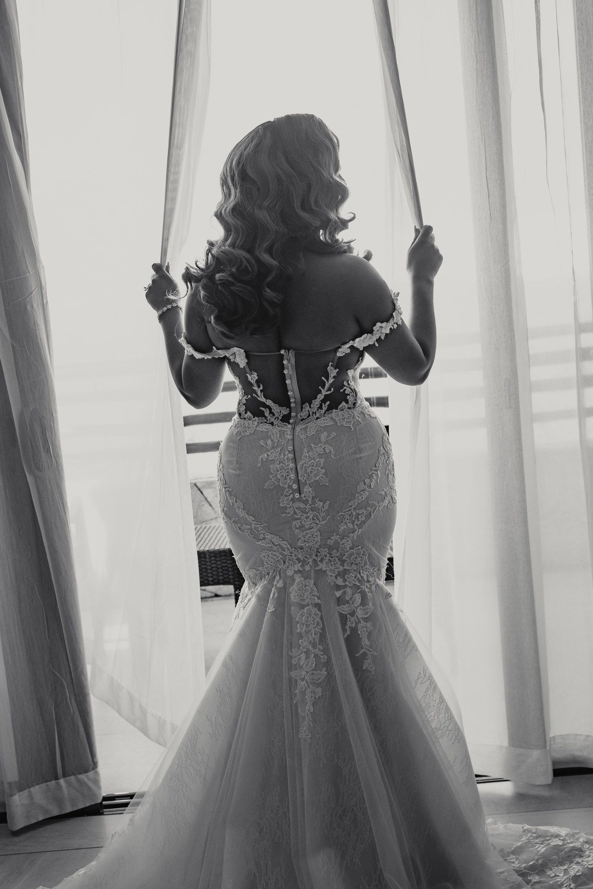 Black and white photo of bride in lace mermaid gown standing at window.