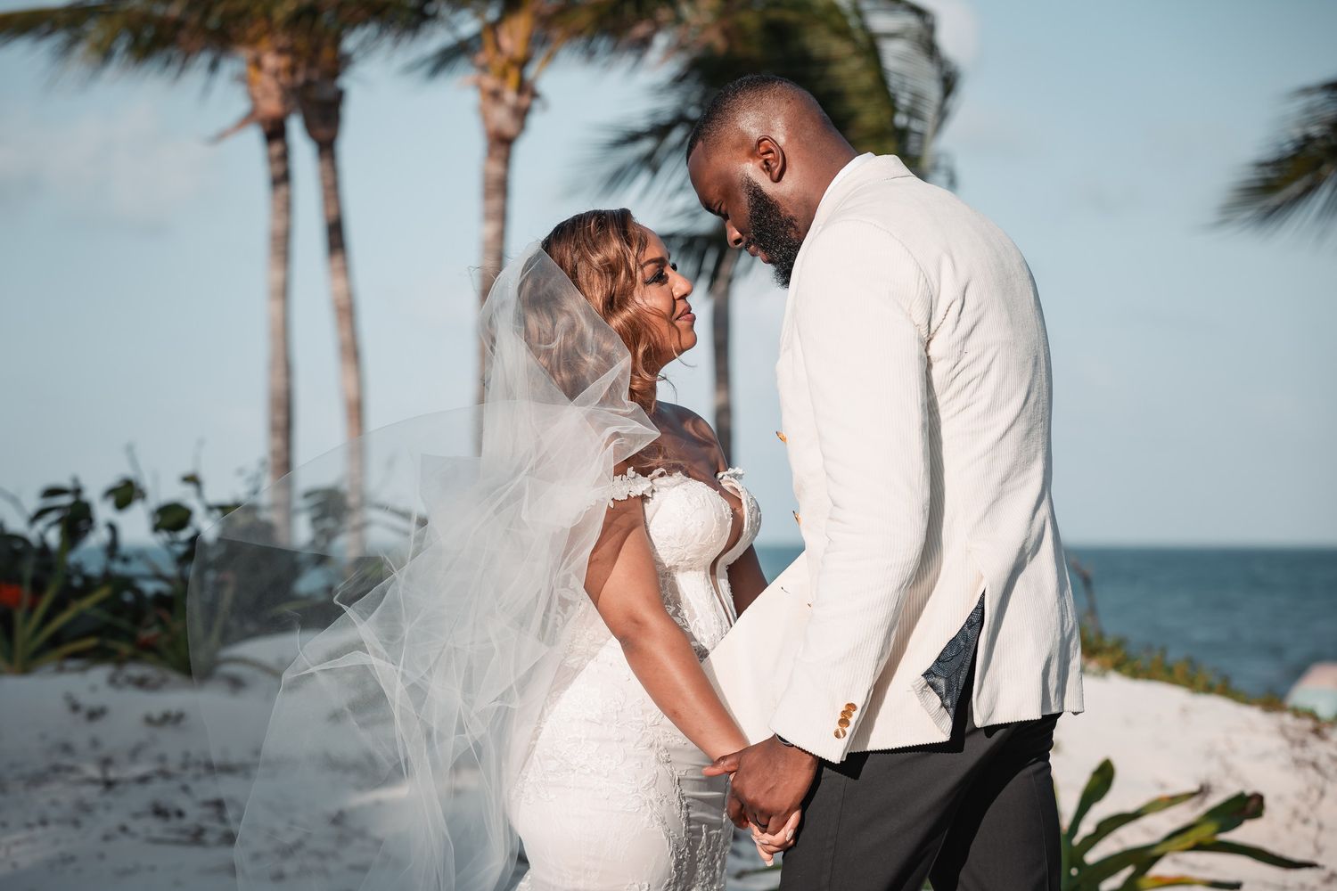 Black couple sharing romantic moment on tropical beach, bride in white gown and veil, groom in white suit.